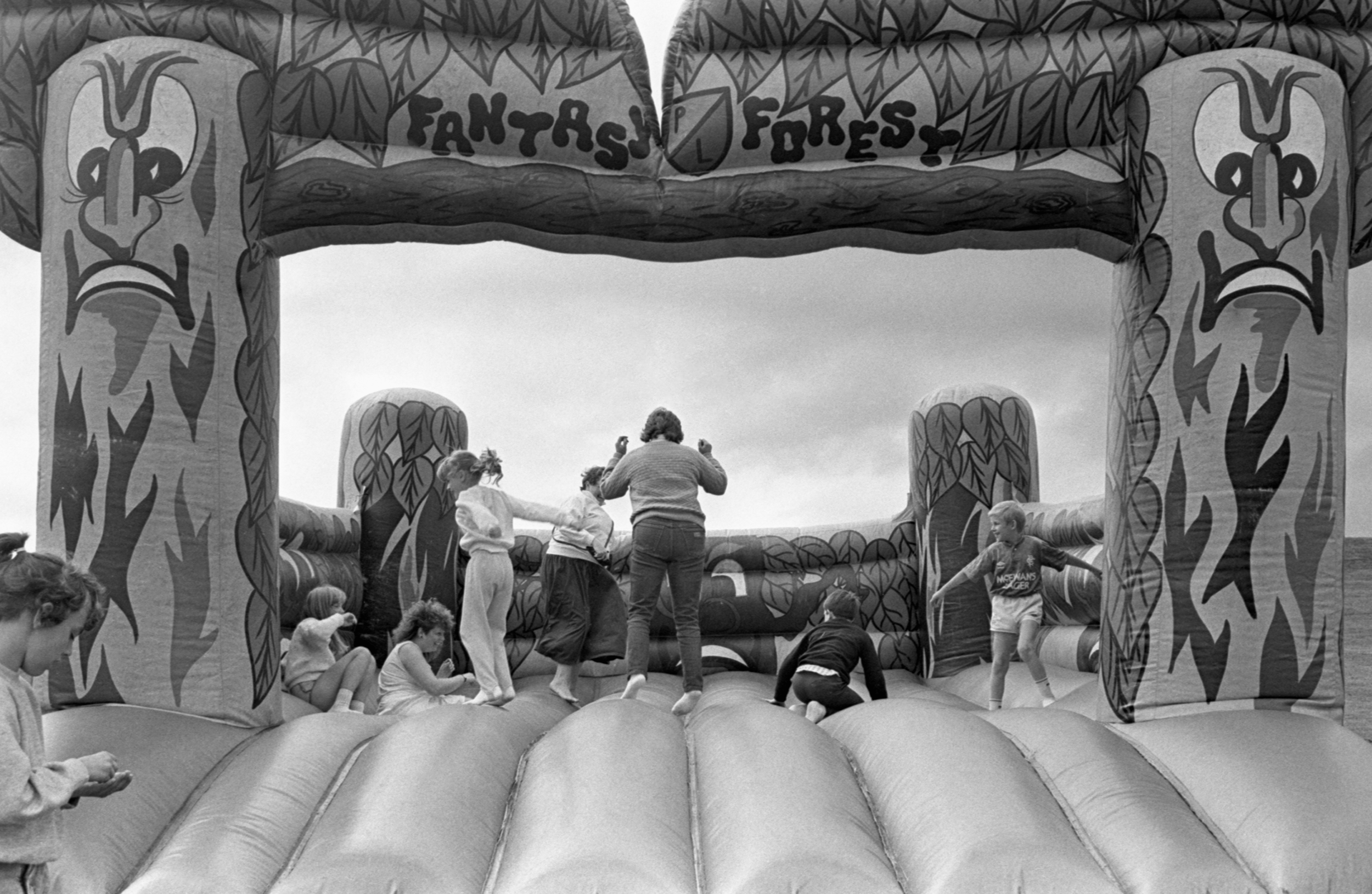 Image description: black and white landscape photograph of children and adults on a FANTASY FOREST bouncy castle. Some are sitting, and some are mid bounce. The bouncy castle has two pillars painted like angry trees. The adults and children are laugh