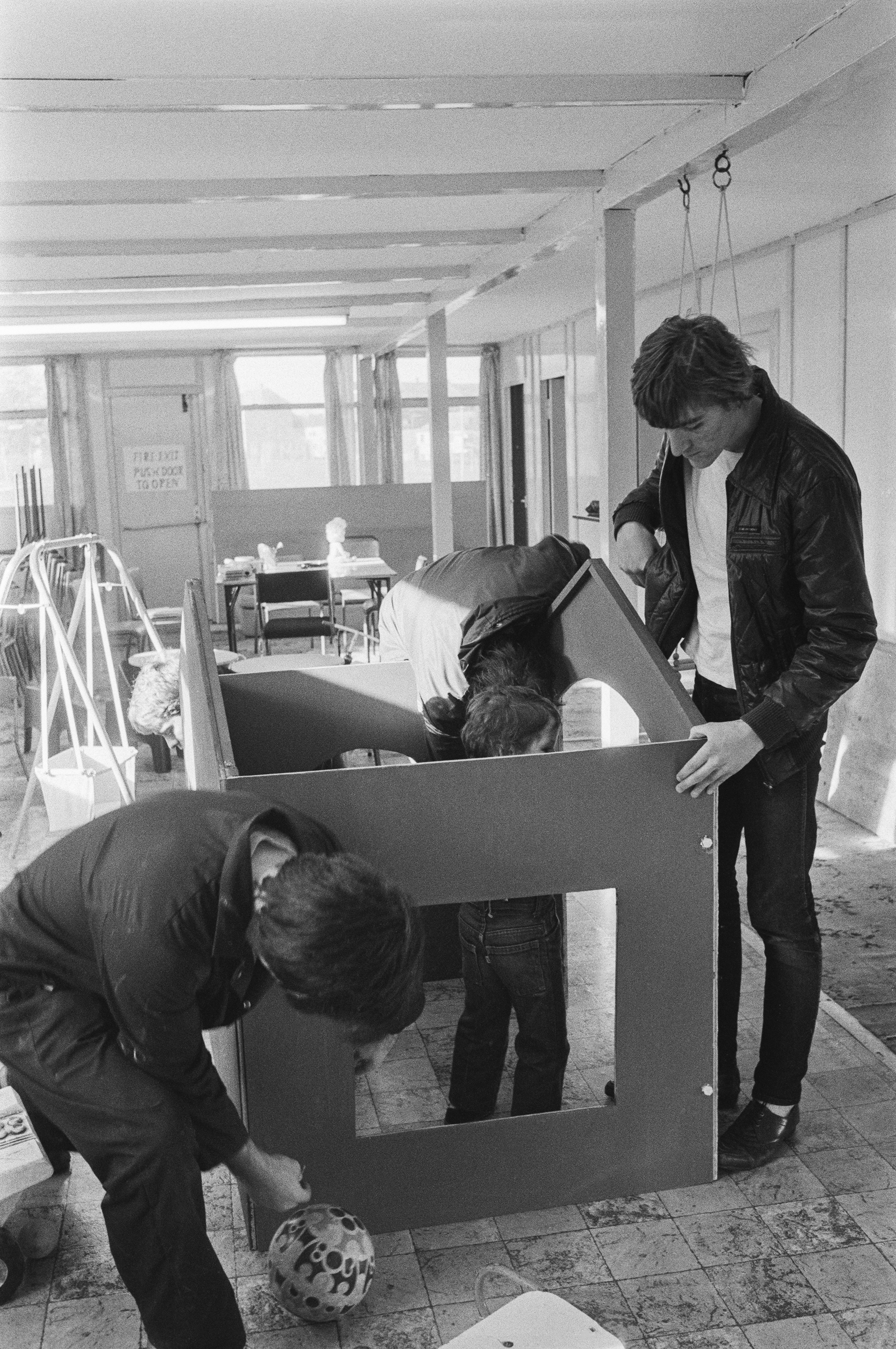 Image description: black and white portrait photograph of three adults and a child constructing a play house. On the left, an adult with light skin, dark hair, and dark overalls bends down inspecting a corner. In the back right, another adult does th