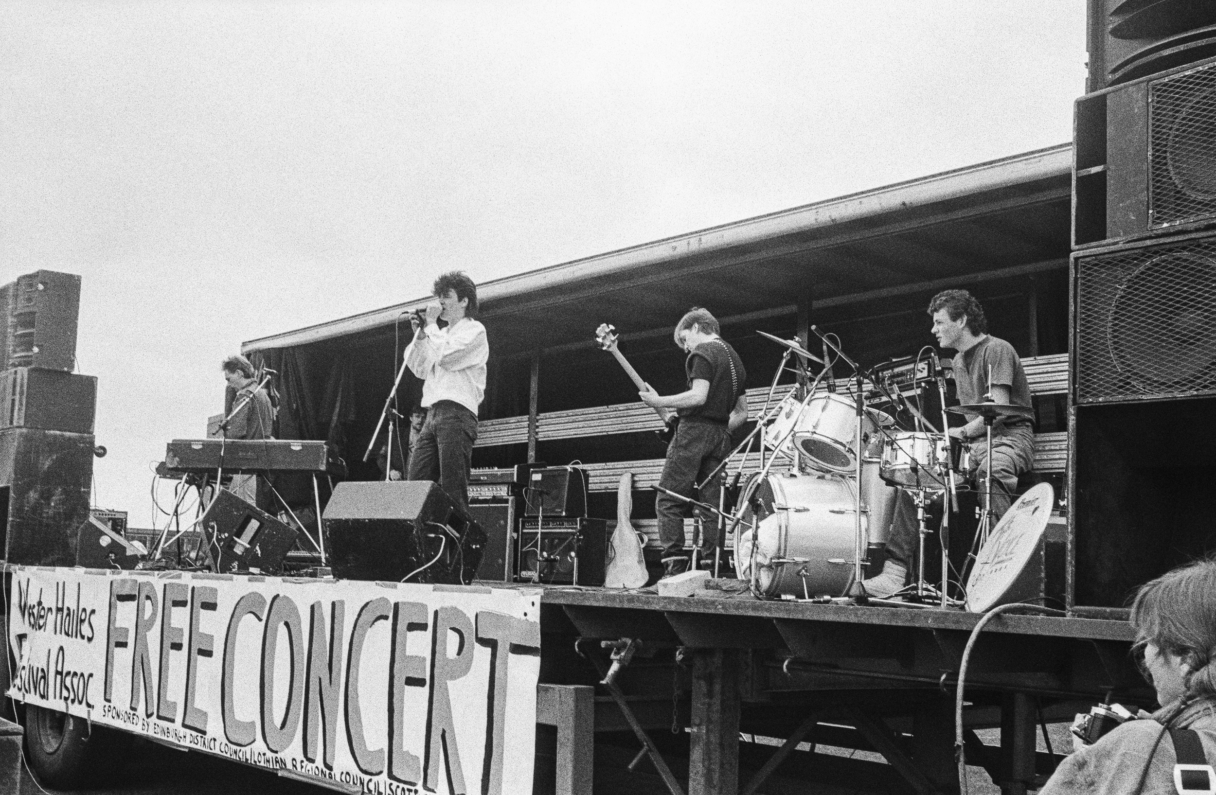 Image description: black and white image of a band peforming onstage. The stage is raised, with speakers on either side and a dark backdrop with silver metallic stripes. A banner hangs from the front reading "WESTER HAILES FESTIVAL ASSOC. FREE CONCER