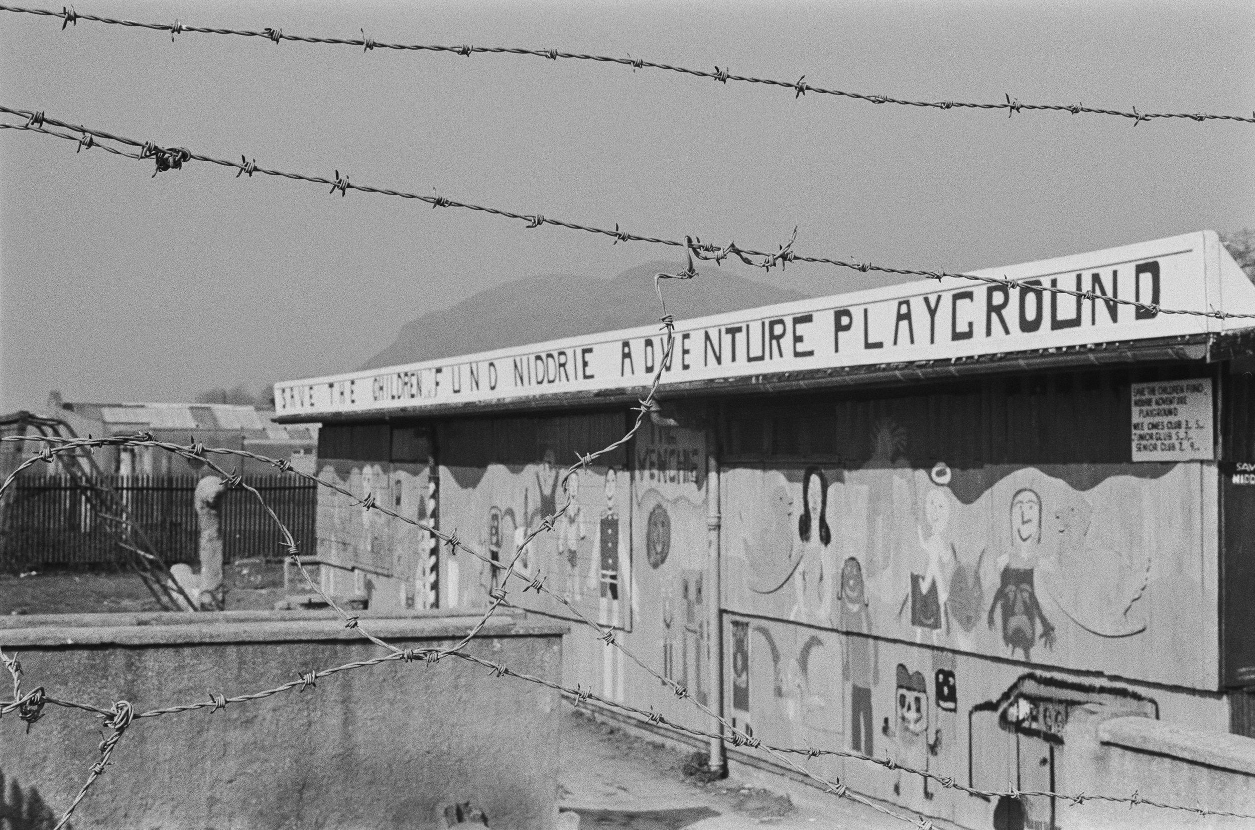 Image description: black and white landscape photograph of the Venchie Young People's Project building behind barbed wire. The building has a mural depicting children smiling. Across the building's top, a sign reads 'SAVE THE CHILDREN FUND NIDDRIE AD