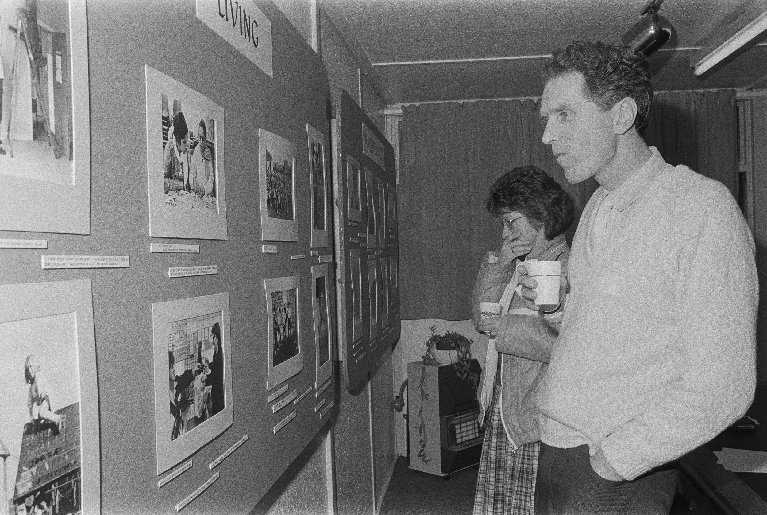 Image description: black and white landscape photograph of two people looking at a wall of photographs in an exhibition. In the foreground, a man with light skin and short medium dark hair wearing a white jumper and grey trousers holds a cup in his r