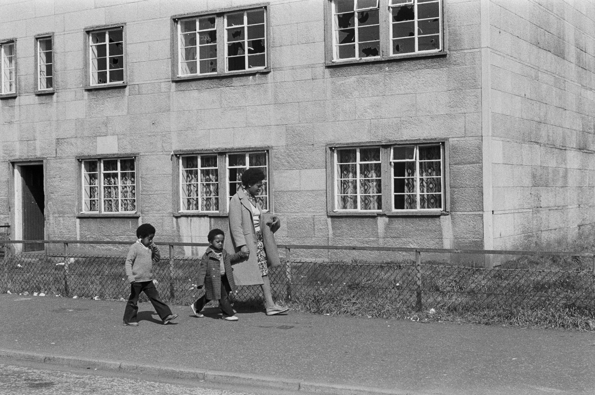 Image description: black and white landscape image of a woman and two children walking down a street in Niddrie. The woman has dark skin and short dark hair, and is wearing a light coloured coat, a patterned dress, and a white pullover jumper. She is