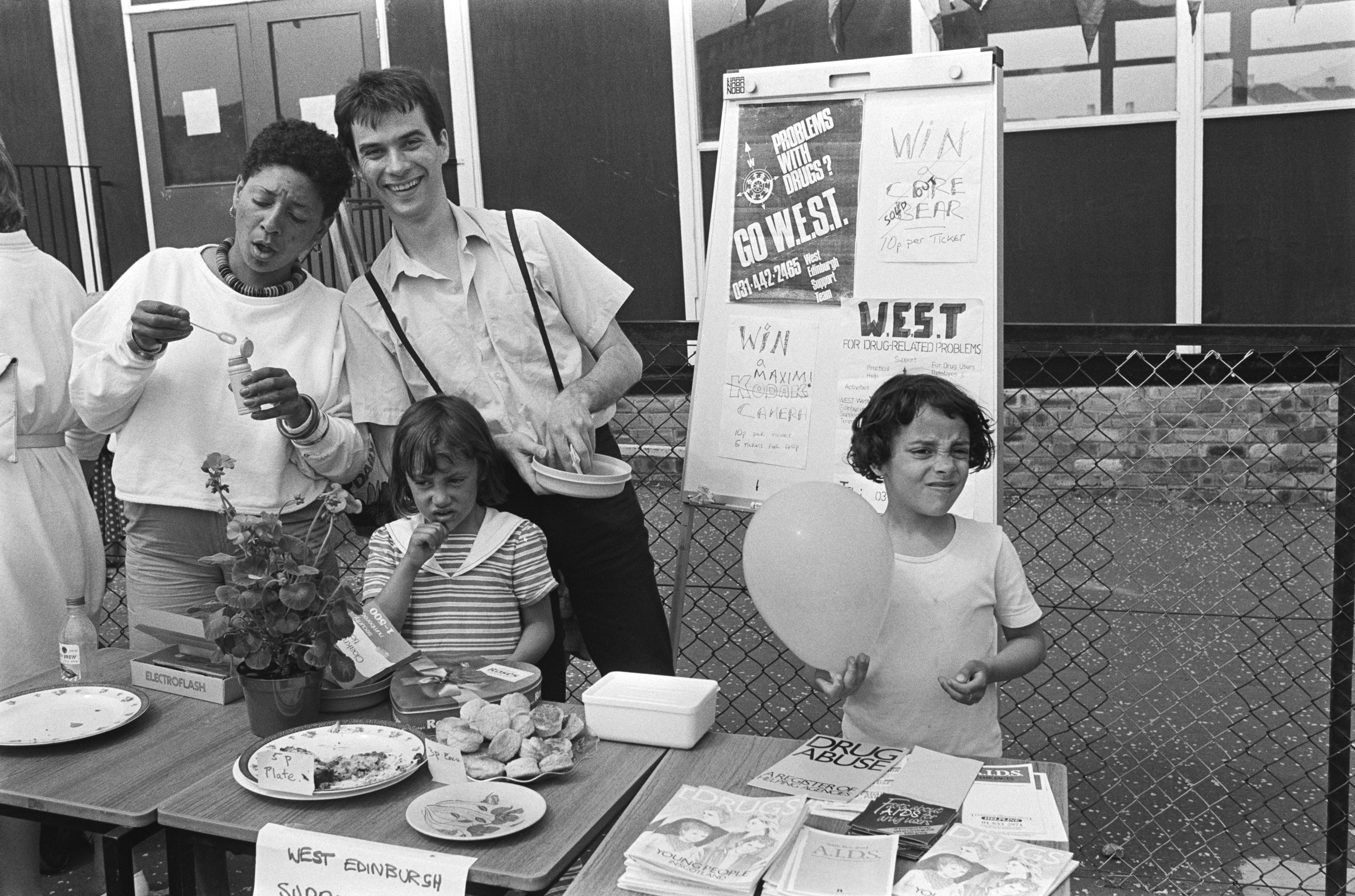 Image description: black and white portrait of two adults and two children standing behind a stall outside in front of a building. The stall is made up of three tables with empty plates, bottles, plates of cakes, a plant, a white plastic tub, a box o
