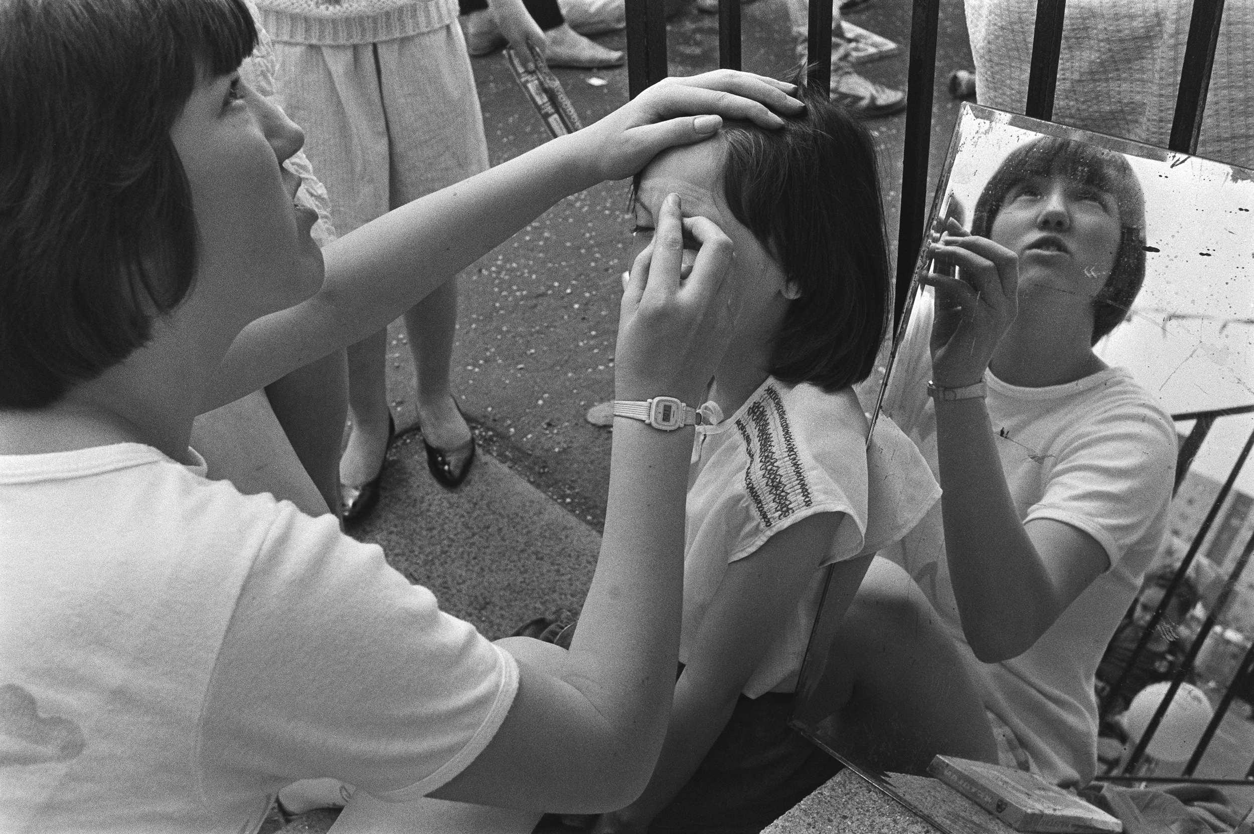 Image Description: black and white portrait of a woman painting a child's face in front of a set of dark railings. The woman on the left of the image has light skin and short dark hair, and looks up towards the top right corner. She is talking to som