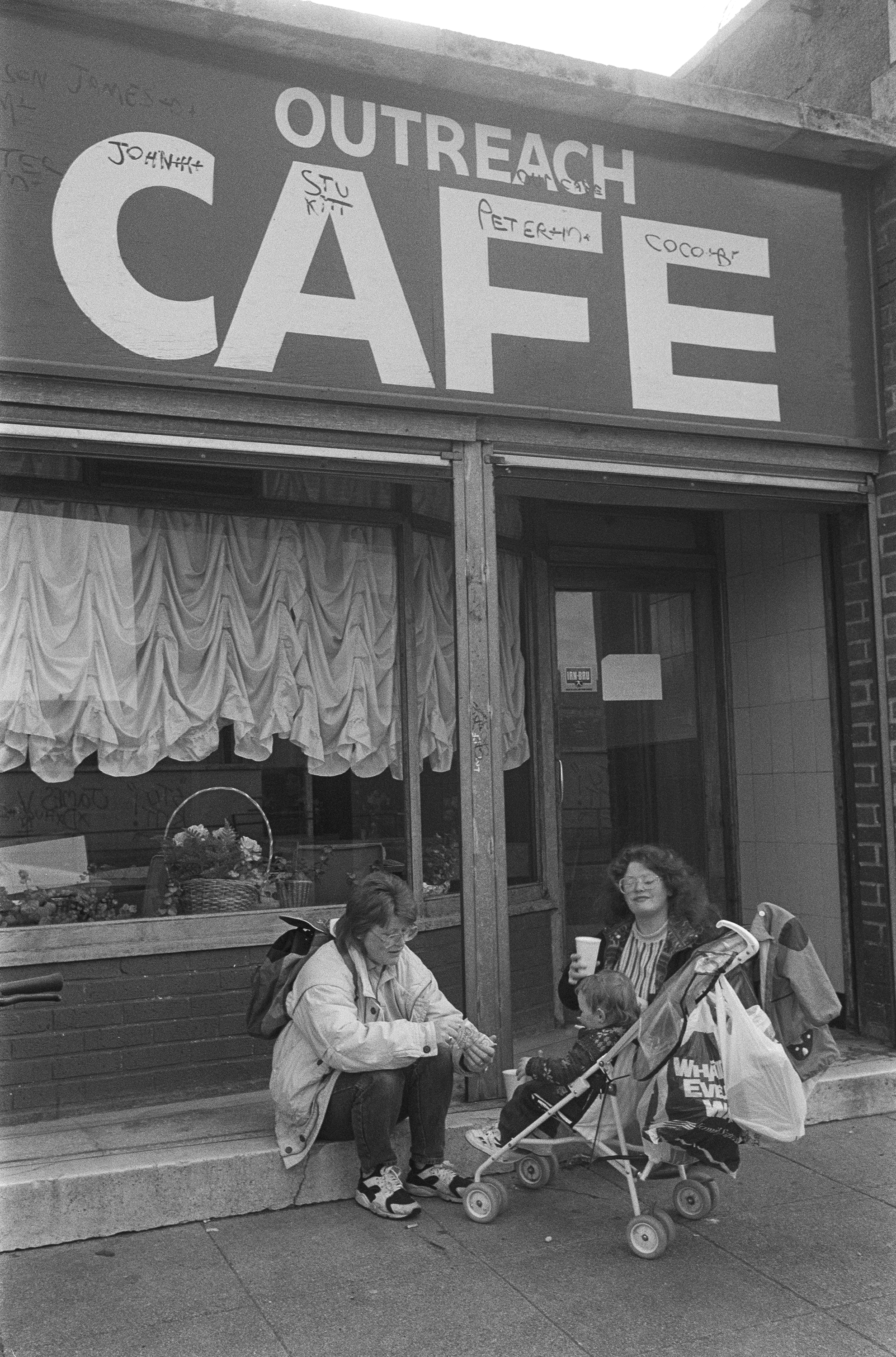 Image Description: A portrait black and white photograph showing the Outreach Cafe. A name has been written inside each letter of the CAFE sign. Two women sit on the steps outside the cafe, one is holding a take out cup and both are smiling at the
