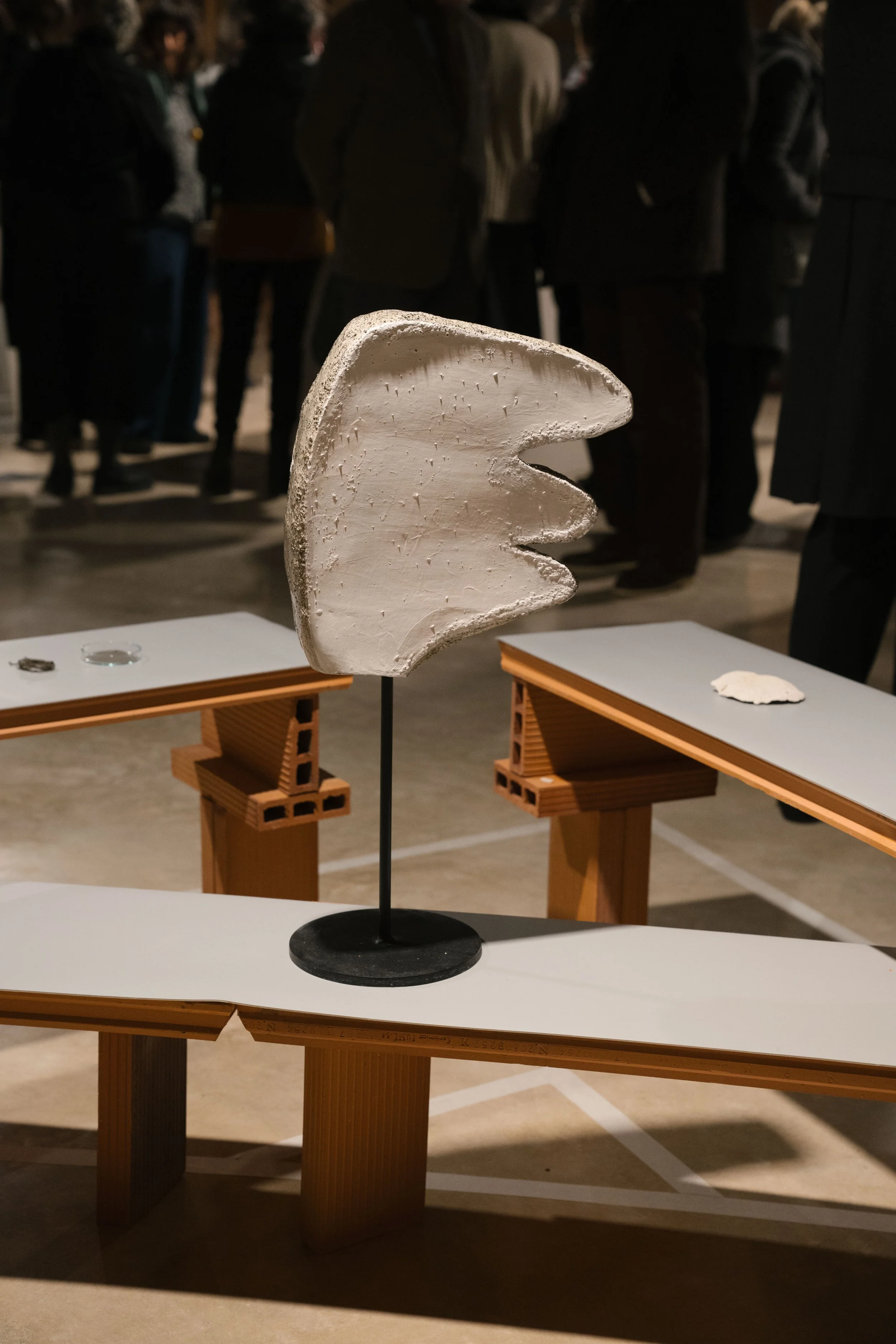 A white ceramic or plaster sculpture with indented lines, mounted on a black stand, displayed on a small white table in an indoor setting with blurred people in the background.