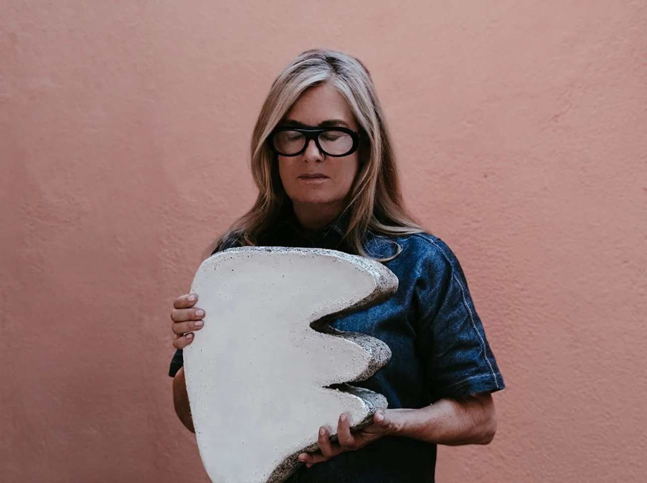 A woman with long blonde hair, wearing glasses and a denim jacket, holding a large, irregularly shaped white piece of art or sculpture against a pink textured wall.
