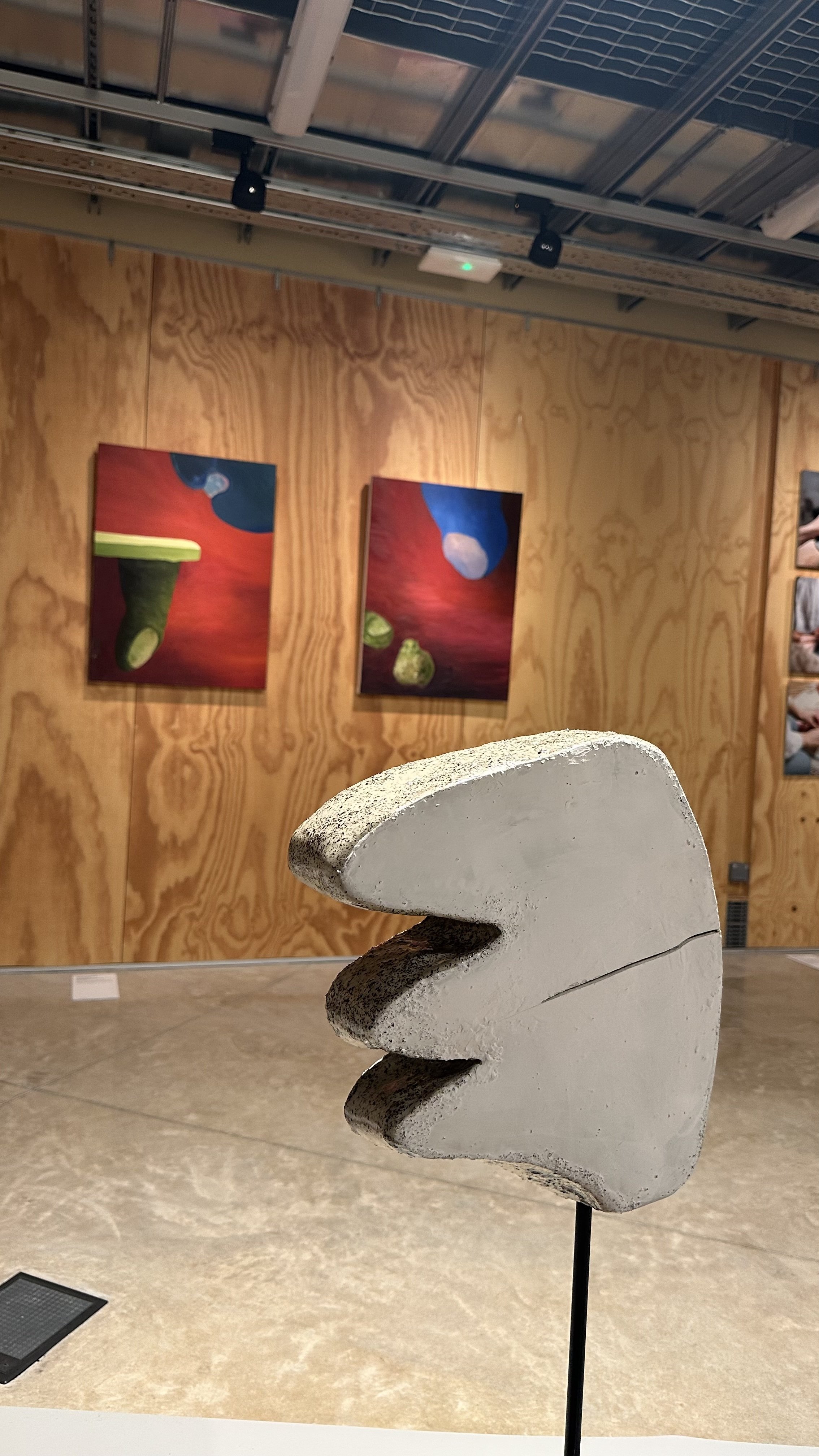 Concrete sculpture resembling Pac-Man with a large open mouth and no eyes, displayed in an art gallery with artwork on wooden-paneled walls.
