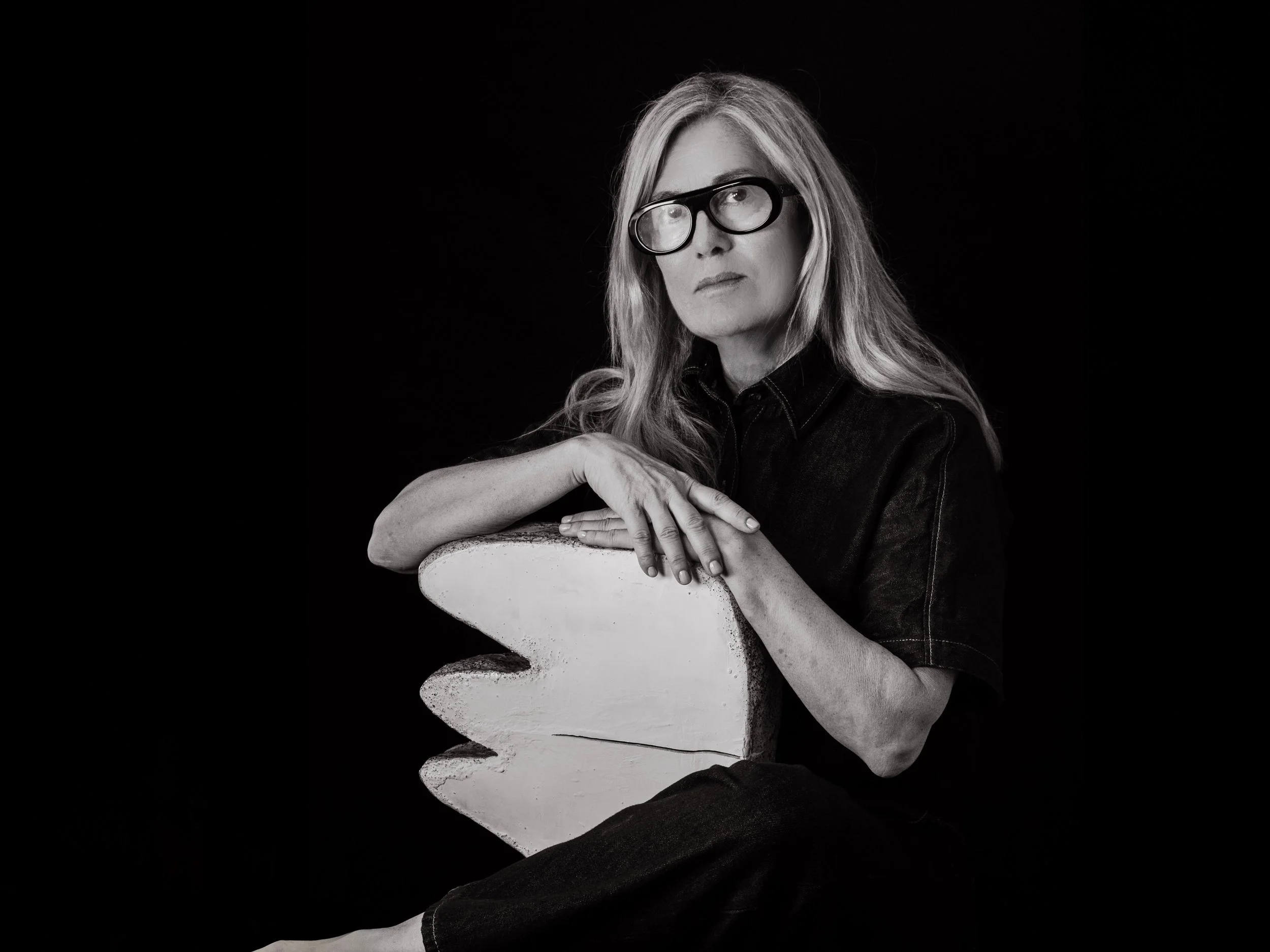 A woman with long hair and glasses, wearing a dark shirt, sitting against a black background, leaning on a painted wooden shark prop.