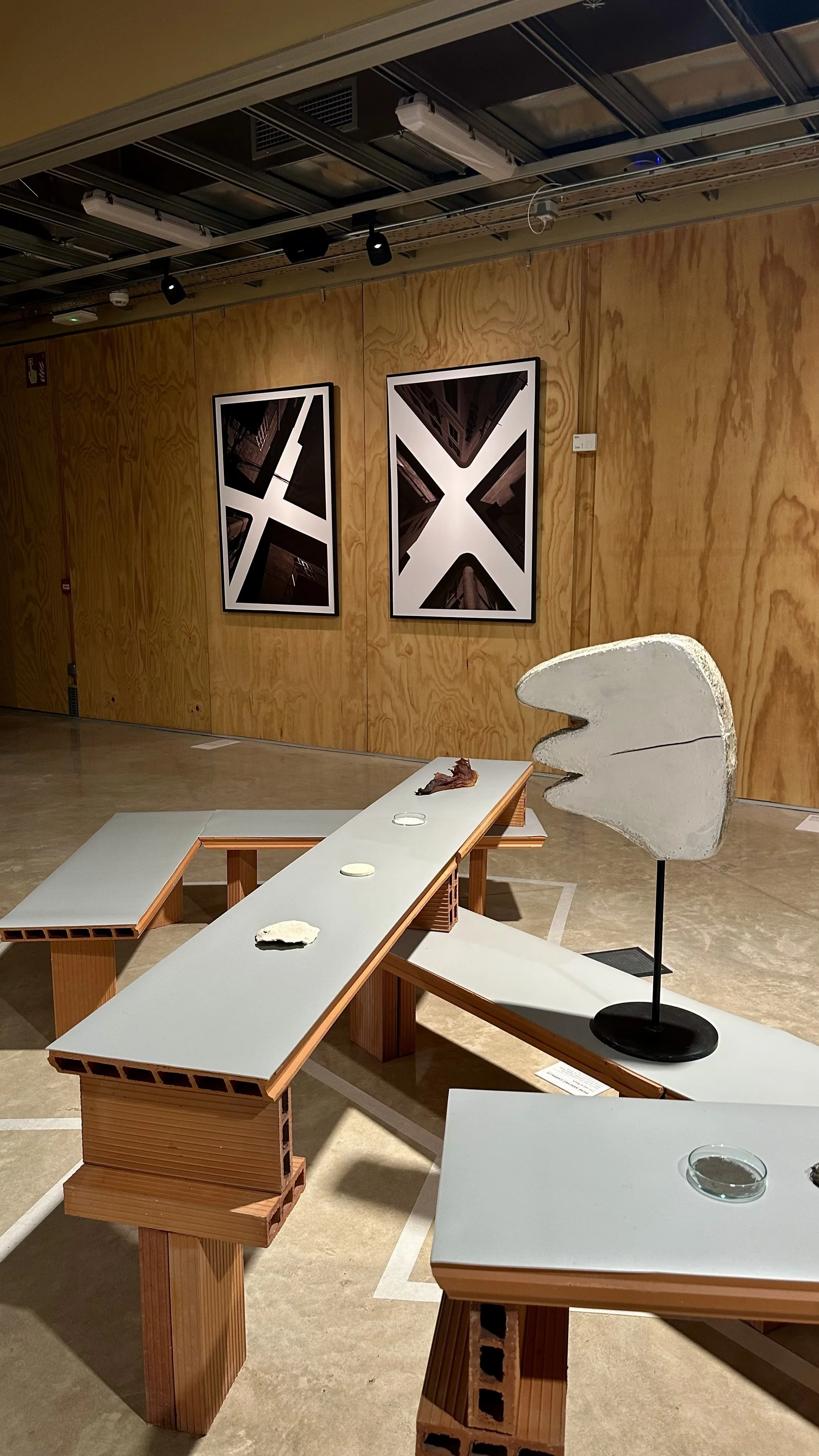 An art exhibition featuring concrete sculptures and black-and-white architectural photographs. The sculptures are displayed on tables made of brick-like structures with white tops, and the photographs are hung on a wooden-paneled wall.