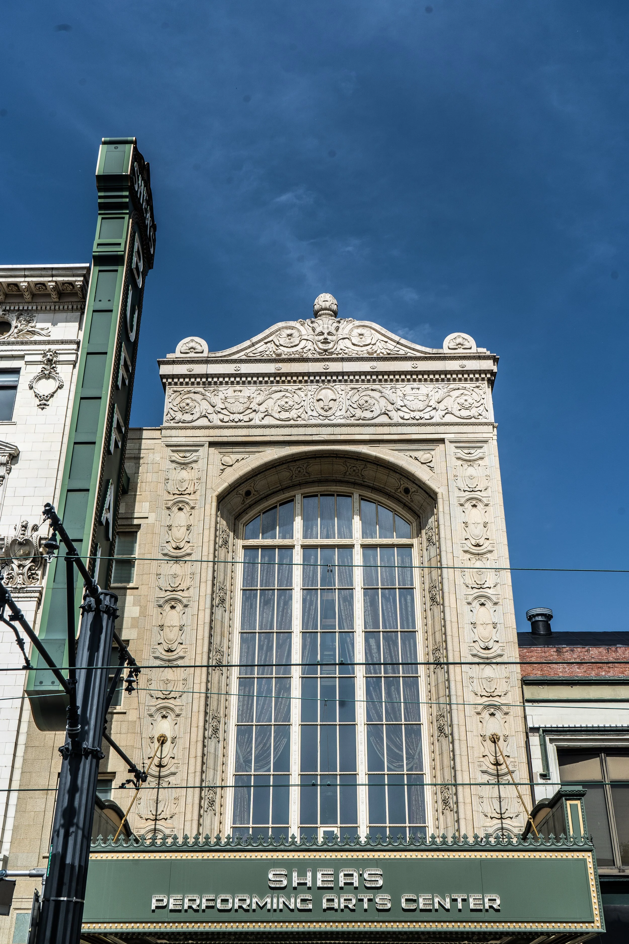 Close-up of the historic Sheas Performing Arts Center building's ornate architectural details with a large window and decorative carvings, under a clear blue sky during daytime.