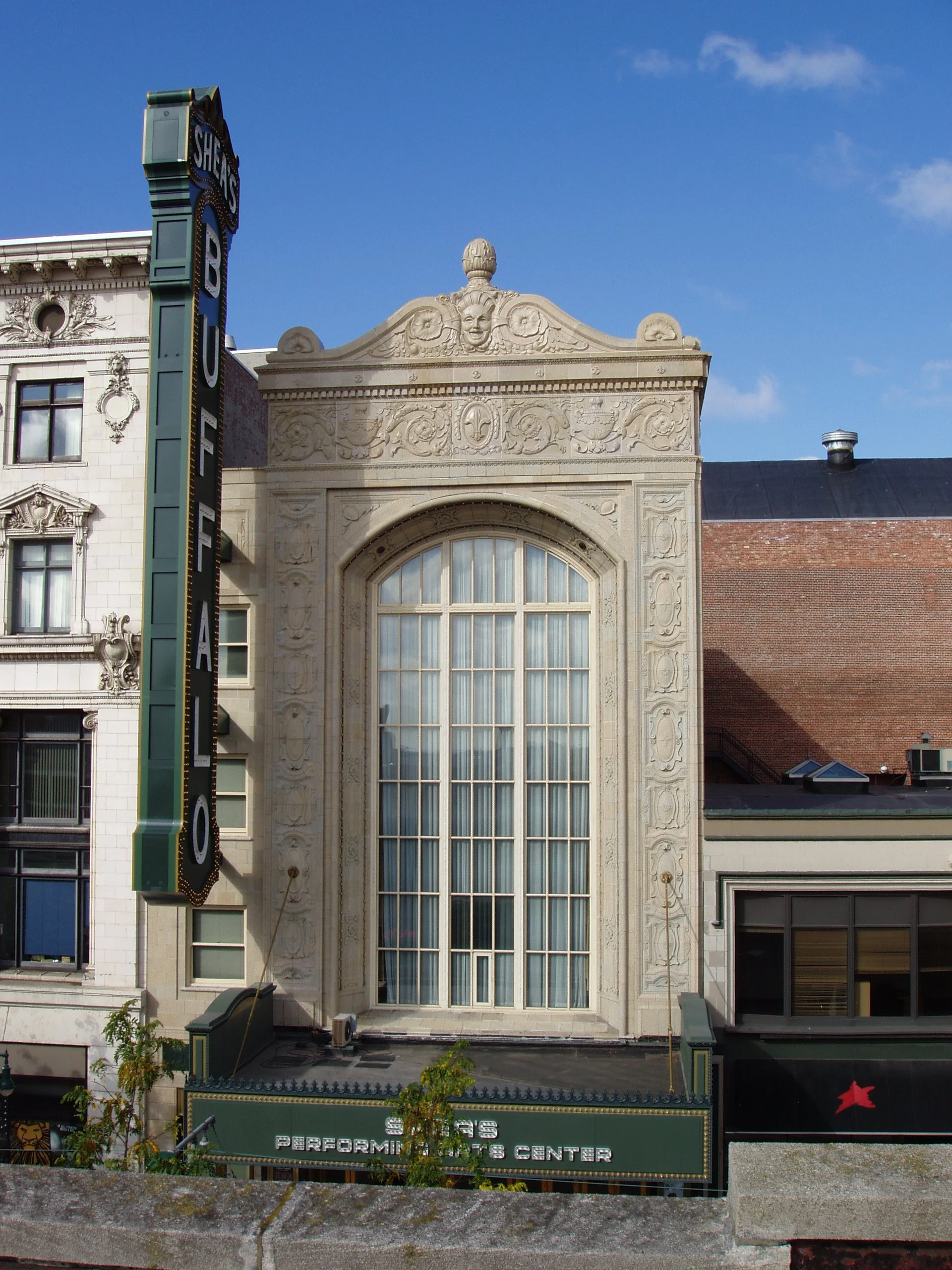 Front view of an ornate building with a large window, decorative stone details, and a sign that reads 'Shea's Performing Arts Center'. A vertical sign on the left reads 'Shea's'.