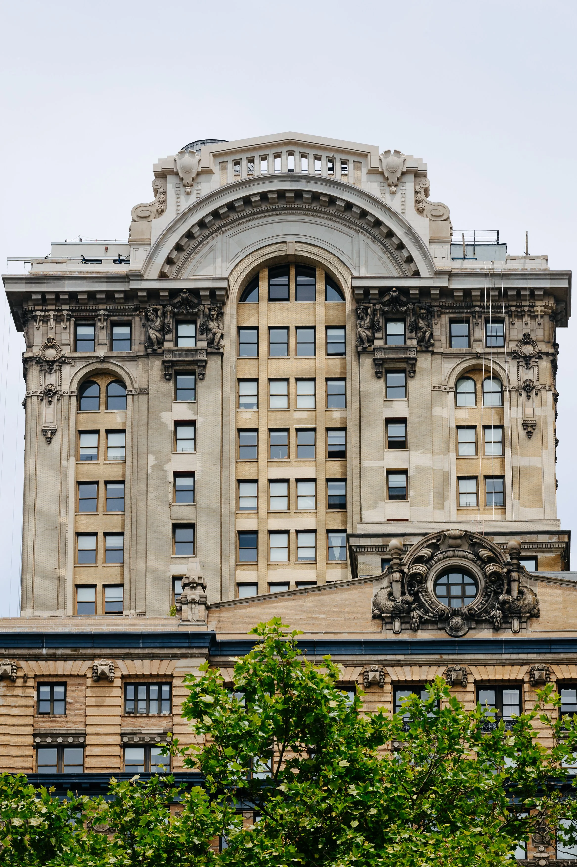 Facade of a historic multi-story building with classical architectural details, sculptures, and decorative elements, partially obscured by leafy green trees.