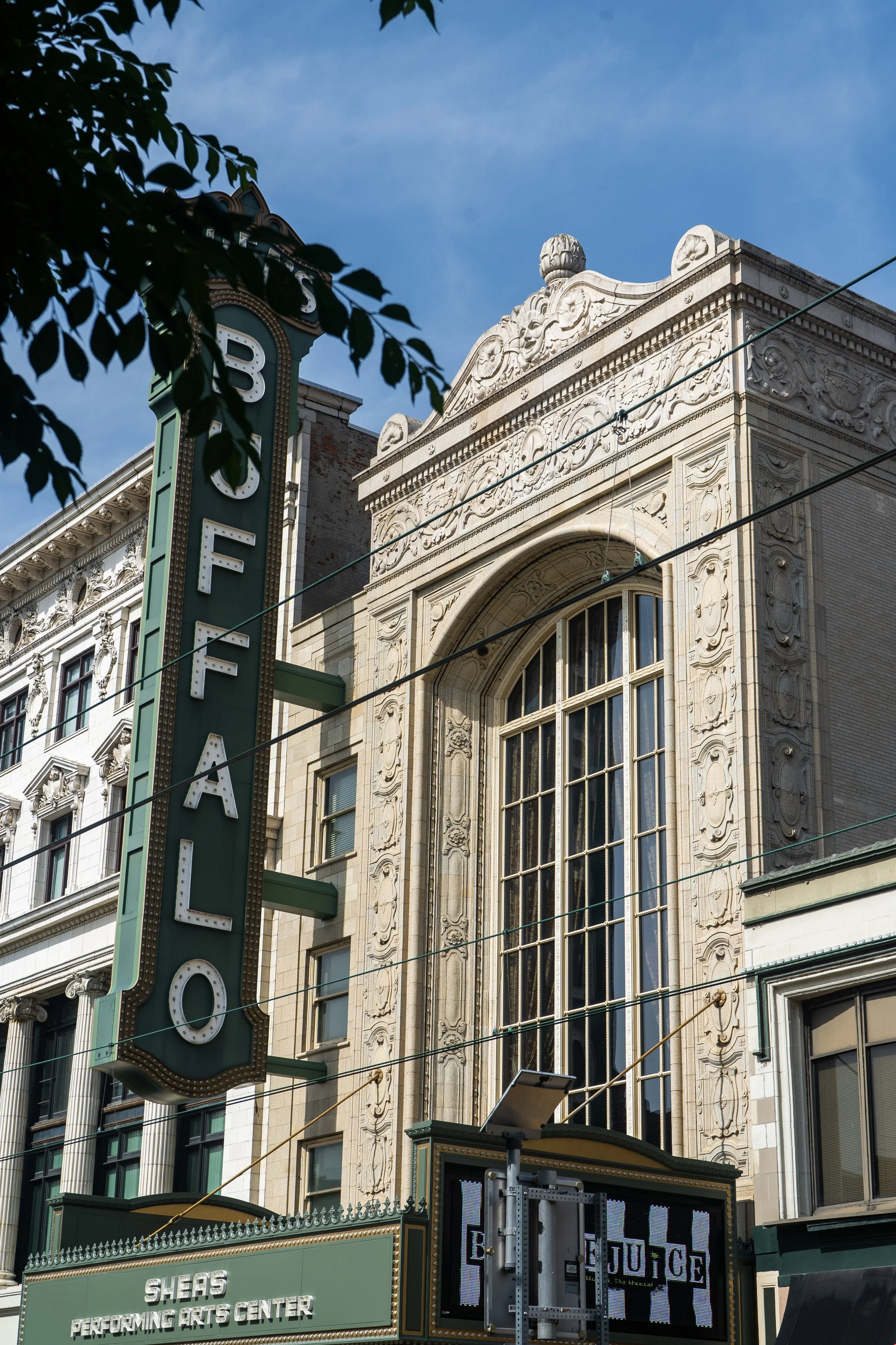 A historic building with ornate architectural details, large arched windows, and a prominent vertical sign reading 'SHEA'S' with additional text about a performing arts center. A multiple-panel electronic sign displays advertisements or information. 