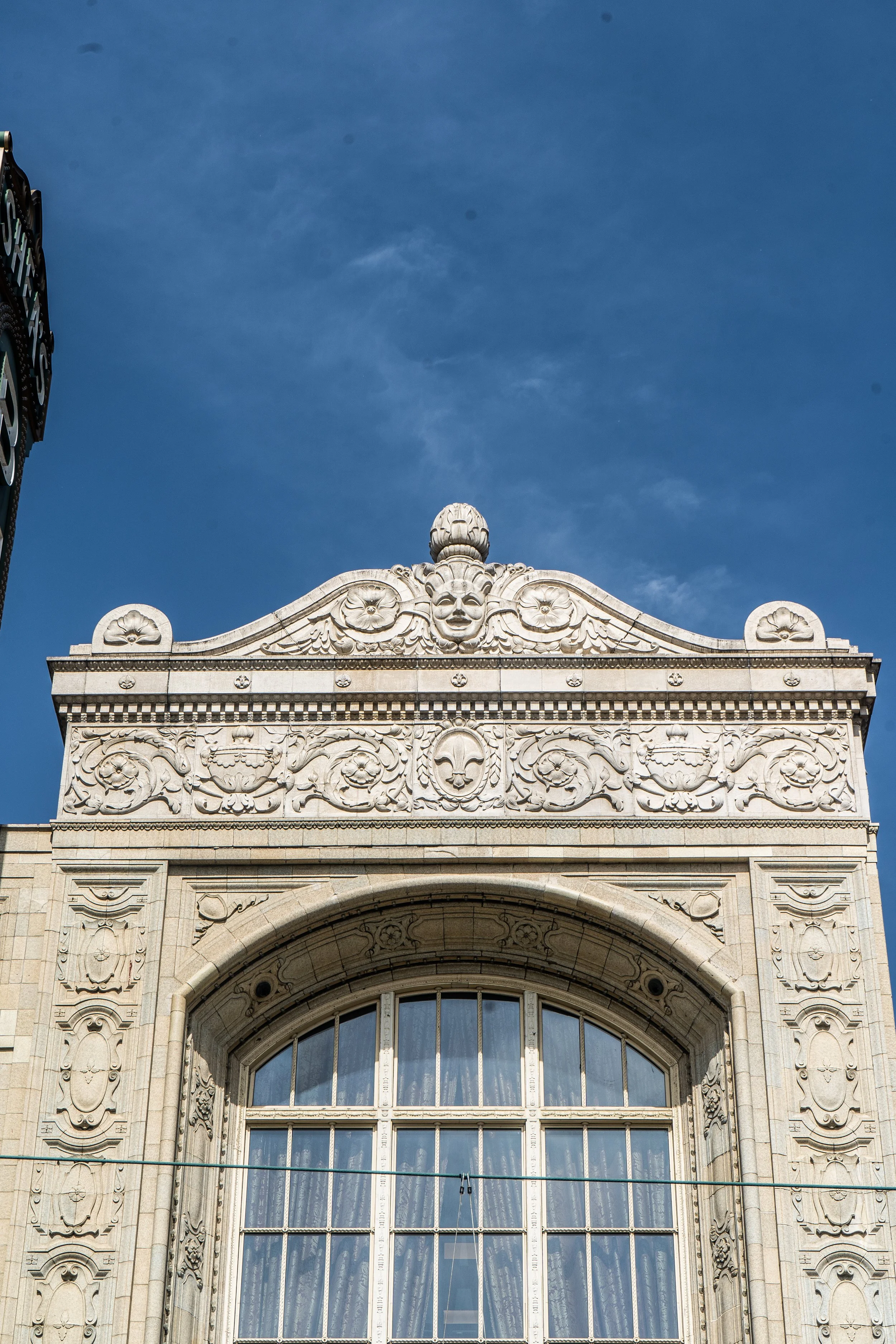 Detailed stone building facade with arched window, decorative carvings, floral motifs, and a carved face with a headdress. Blue sky in the background.