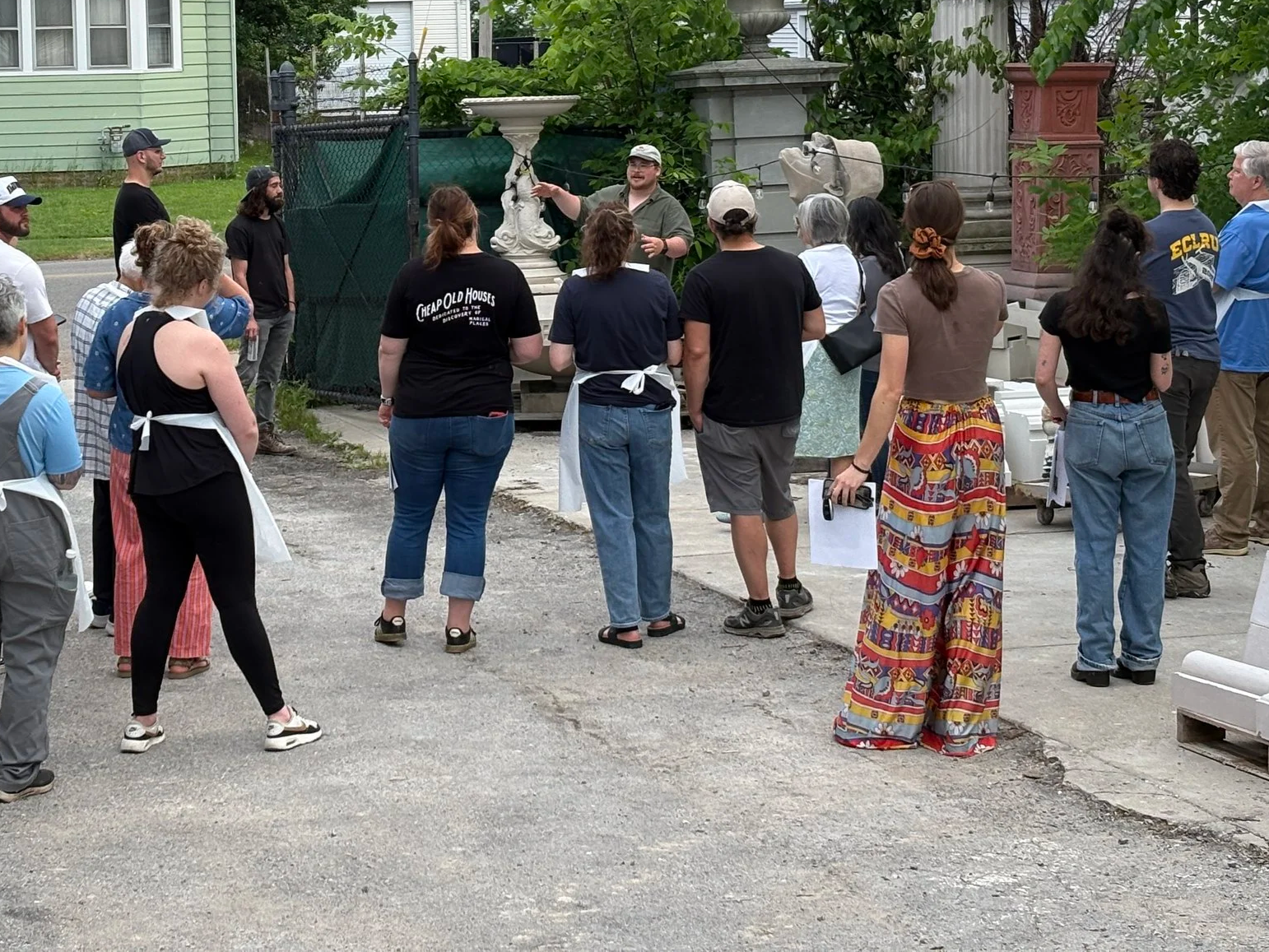 A group of people gathered outdoors around a tour guide who is speaking. The group includes men and women of various ages, some wearing aprons and casual clothing. The setting appears to be an outdoor area with decorative items and greenery in the background.