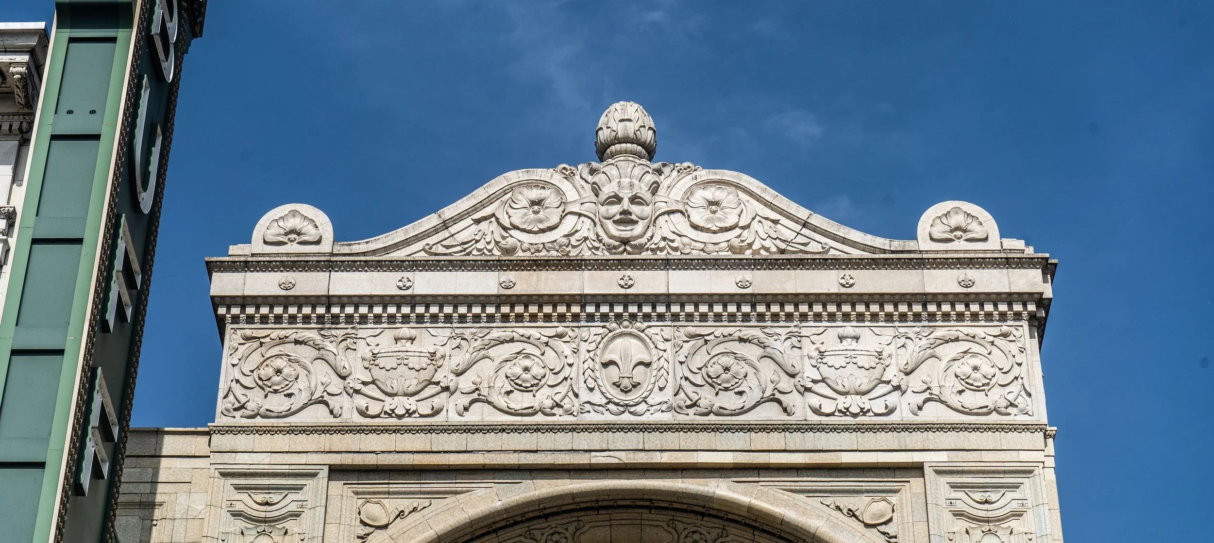 Decorative stone building facade with an ornate carved design, a central smiling face, and a large urn on top, against a blue sky.