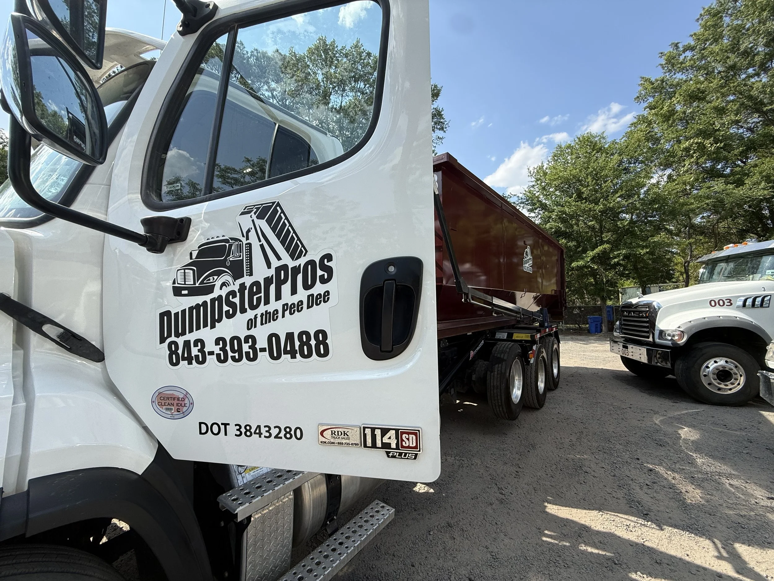 A white DumpsterPros truck with a maroon cargo container is parked outdoors under a blue sky with some clouds, surrounded by green trees and other vehicles.