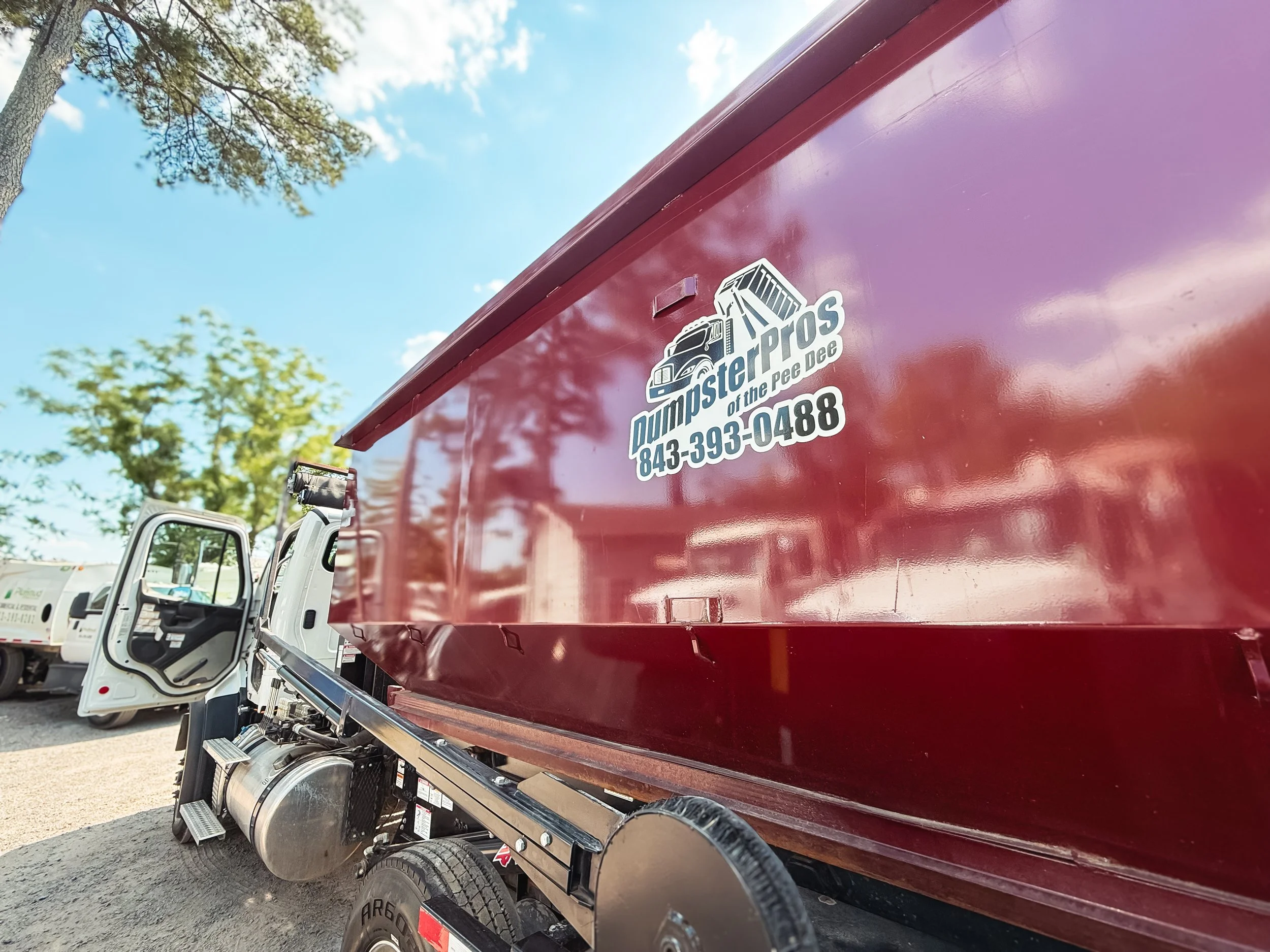 Red commercial trailer attached to a truck, with a white logo and text for Dumpster Pros of the Pee Dee, including a phone number, visible near a tree and blue sky.