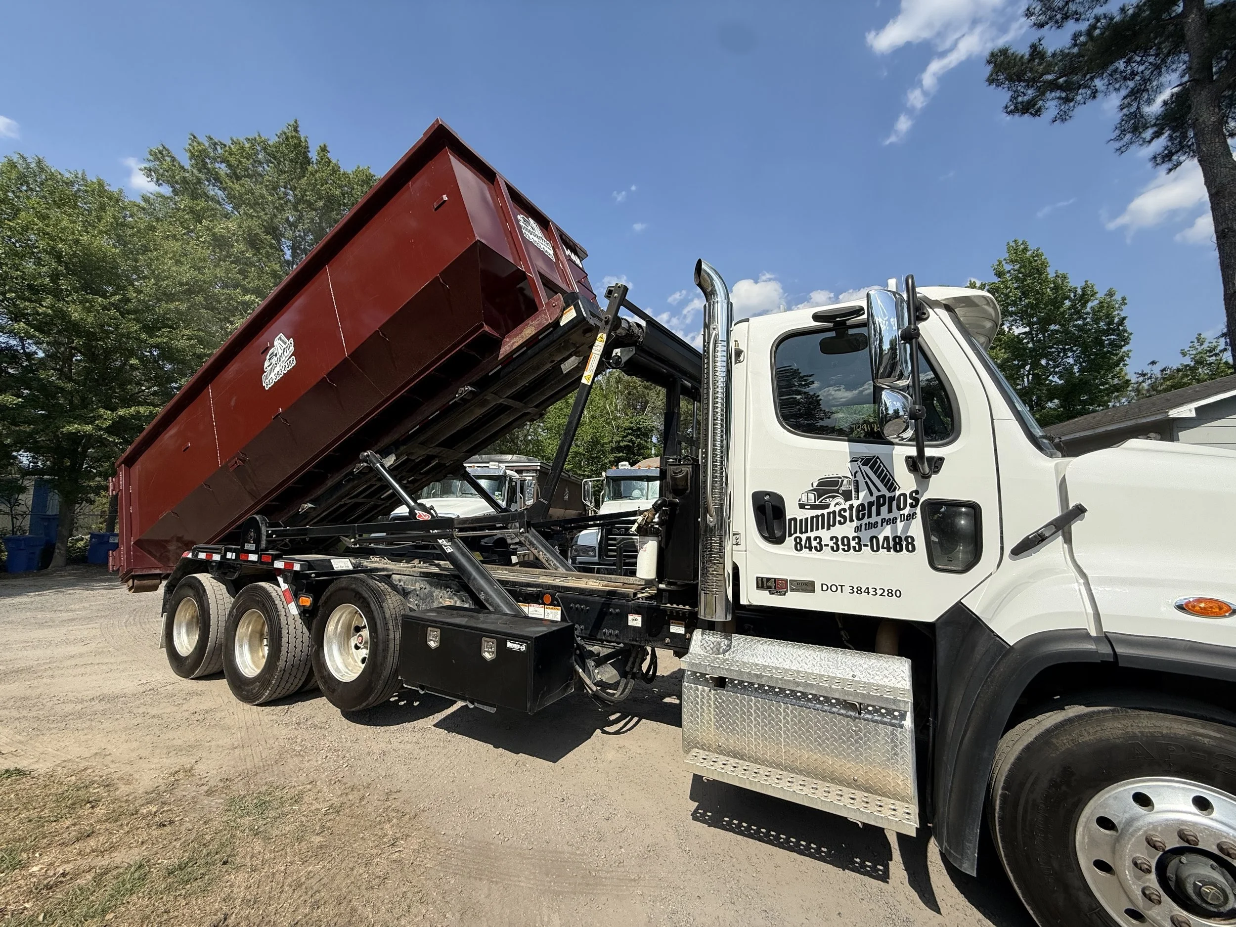A white dumpster truck with a red dumpster bed partially raised, parked on a dirt lot with green trees and blue sky in the background.