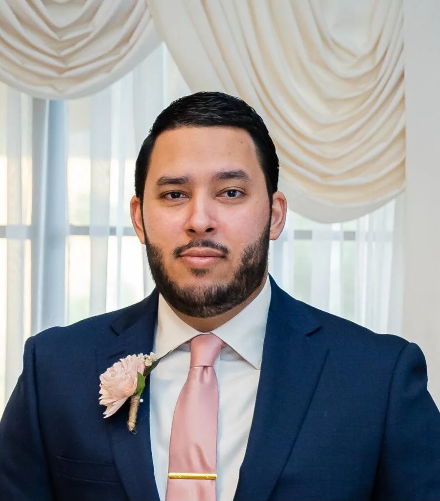 A man in a navy suit, white shirt, and pale pink tie stands indoors in front of a window with draped curtains. He has short dark hair and a beard. He wears a pink flower boutonniere on his left lapel.