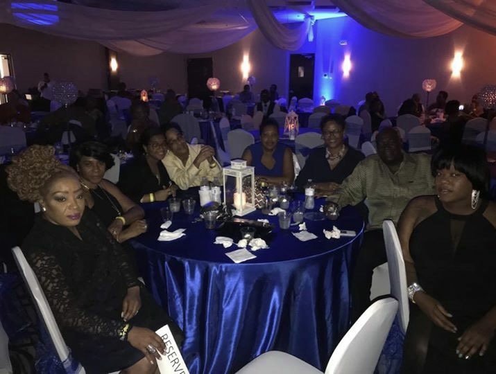 A group of women sitting around a decorated table at a formal event or banquet in a dimly lit venue with blue lighting and draped ceiling.