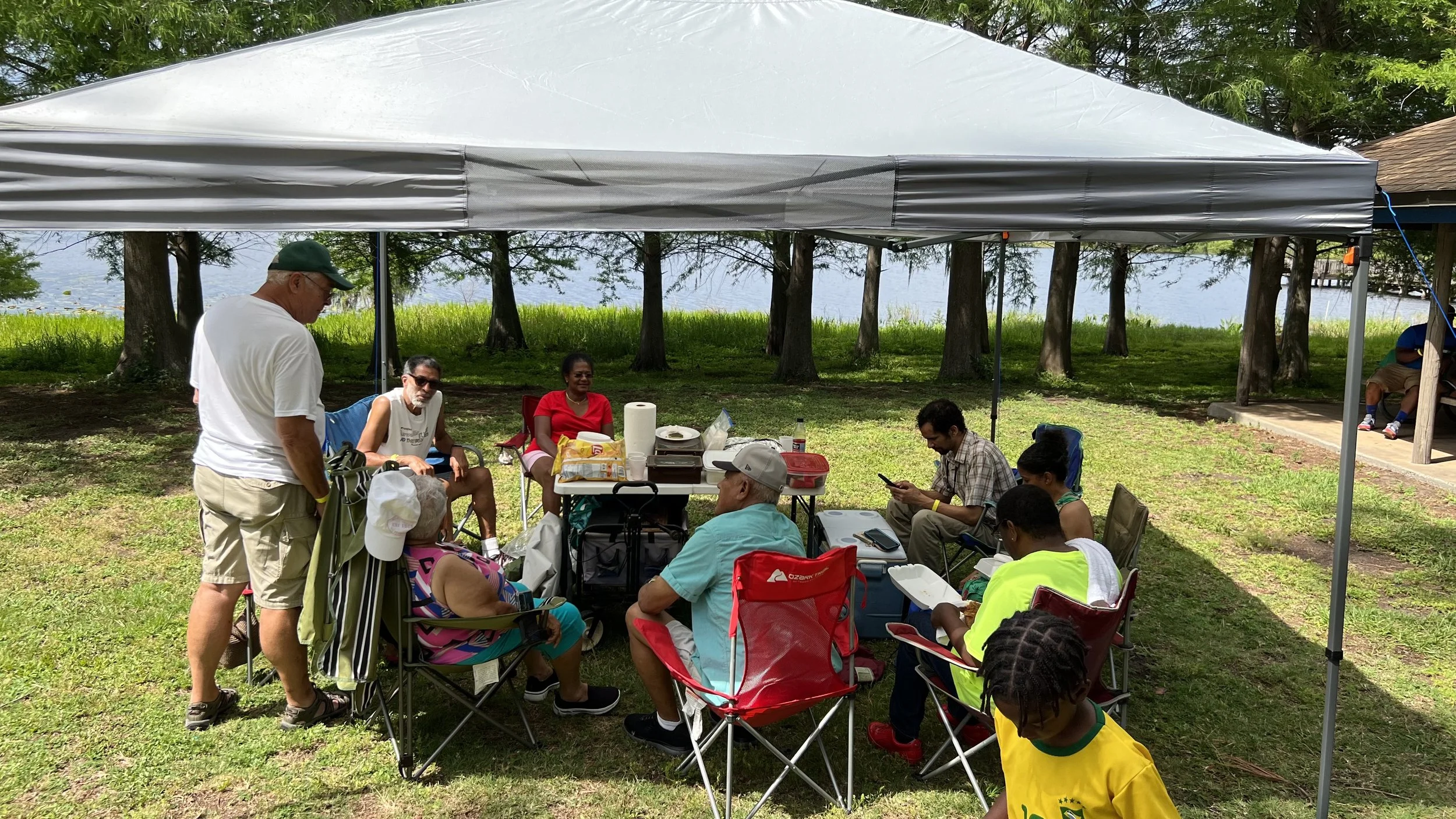 Group of people sitting and standing under a large canopy at a park near water, with trees and grassy area in the background, having a picnic or gathering.