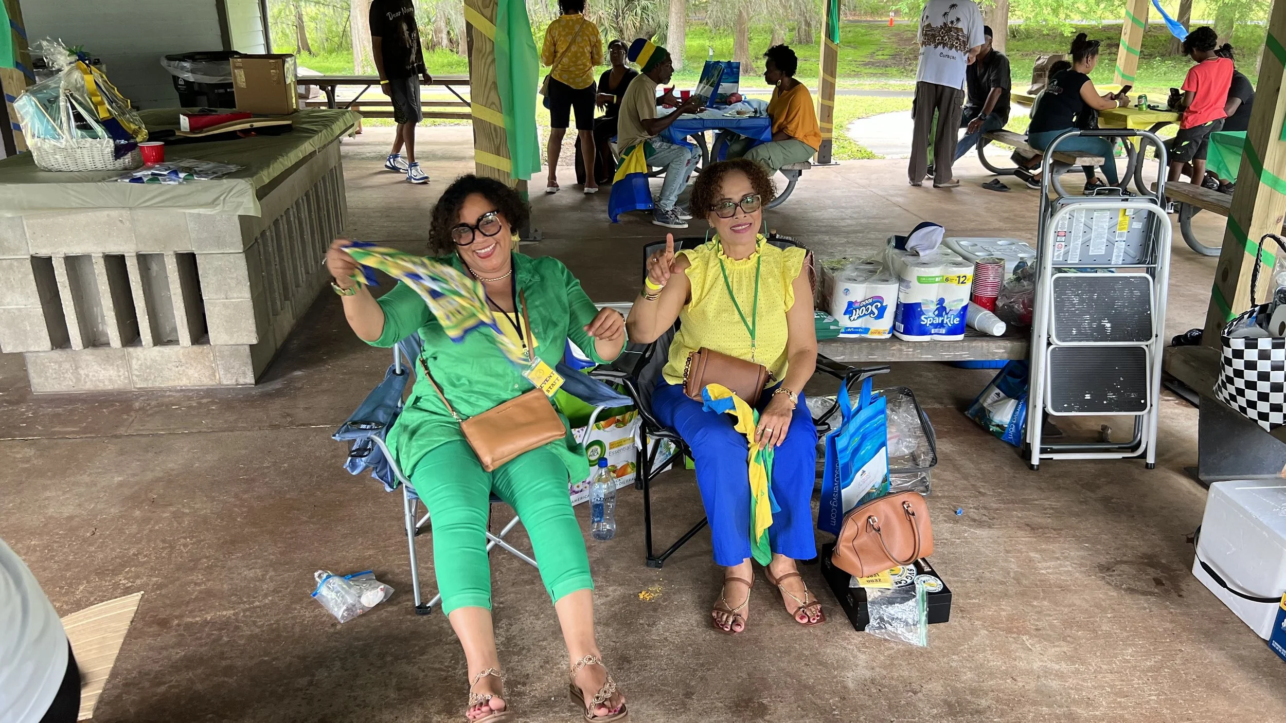Two women sitting on portable chairs at an outdoor gathering, smiling and holding colorful flags or napkins. Behind them are tables with supplies like paper towels, bottled water, and other items. In the background, several people are engaged in vari