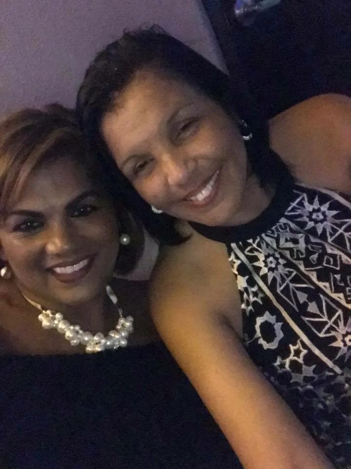 Two women taking a selfie together, smiling, wearing elegant dresses and pearl jewelry in a dimly lit setting.