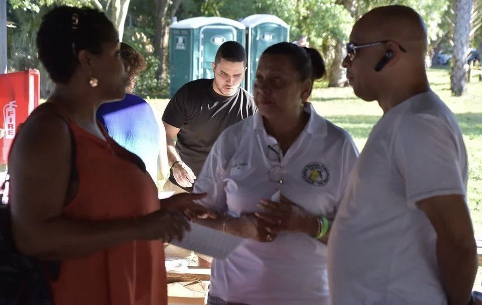 Group of four people outdoors, two women and two men, engaged in conversation. The woman on the left holds papers, and the woman in the middle wears a white polo with a logo. Portable toilets are in the background.