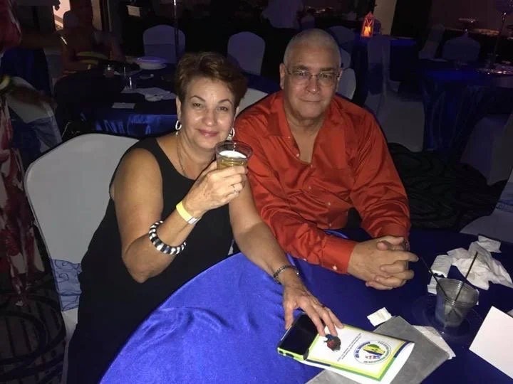 A woman and a man sitting at a table in a dimly lit event or banquet hall. The woman is holding a drink and smiling, while the man is sitting beside her. The table has a blue tablecloth, a menu or program, and a drink with a straw.
