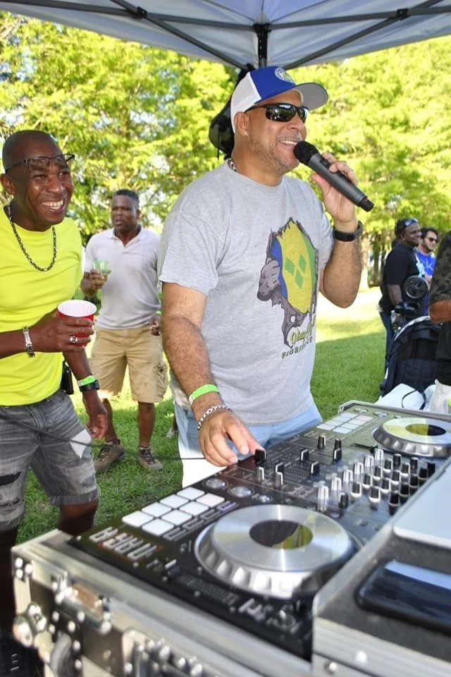 A man in a gray t-shirt, sunglasses, and a blue and white cap is speaking into a microphone while DJing at an outdoor event. Other people are standing around and enjoying the gathering in a park with green trees.