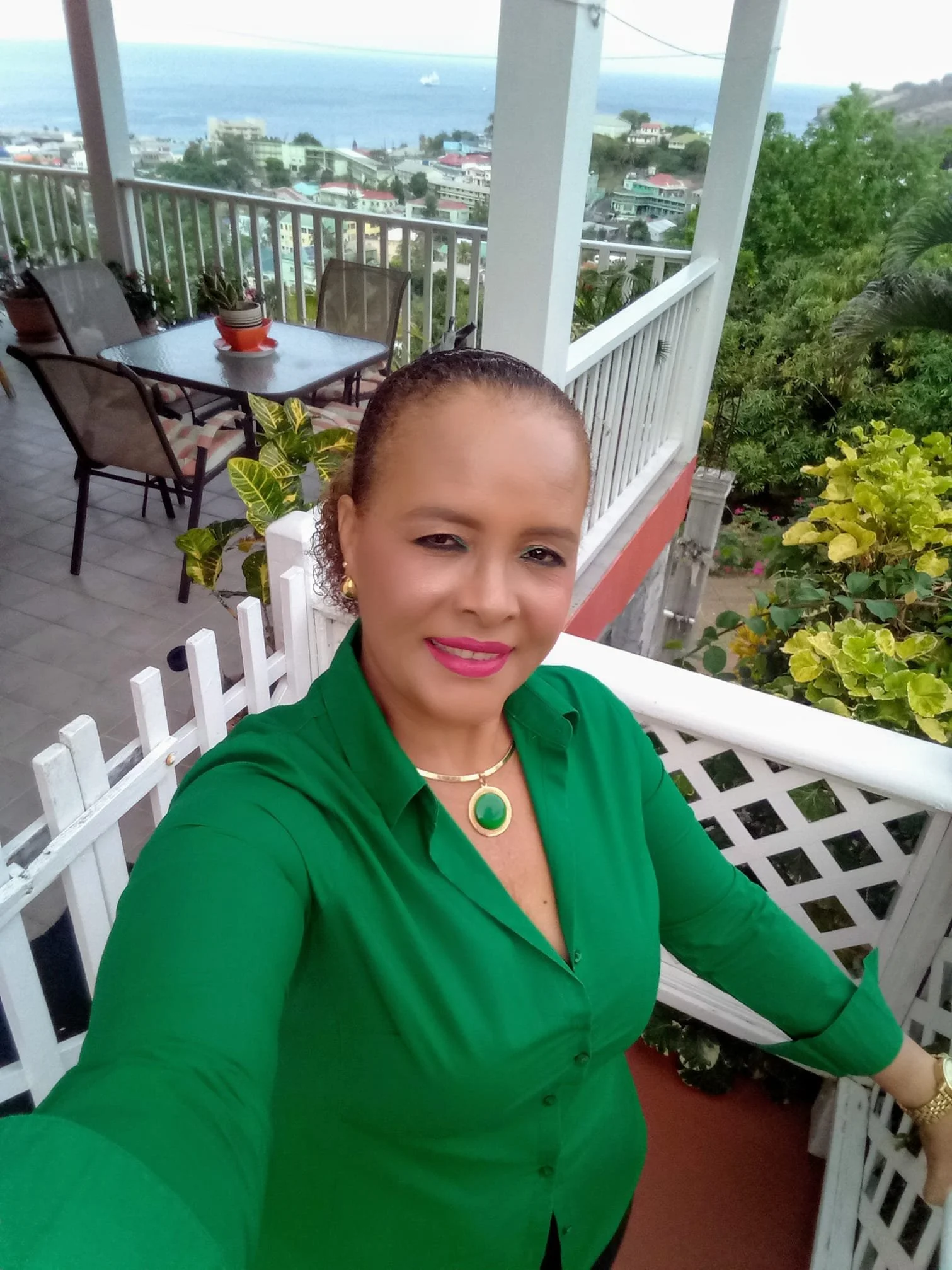 A woman taking a selfie on a balcony overlooking a beach and a coastal town. She is wearing a green blouse, gold jewelry, and has short curly hair.