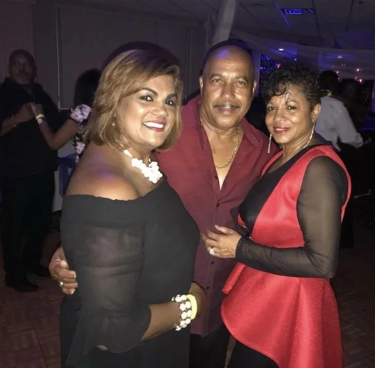 Three people, two women and one man, are smiling and posing for a photo at a party or event, with other attendees in the background.