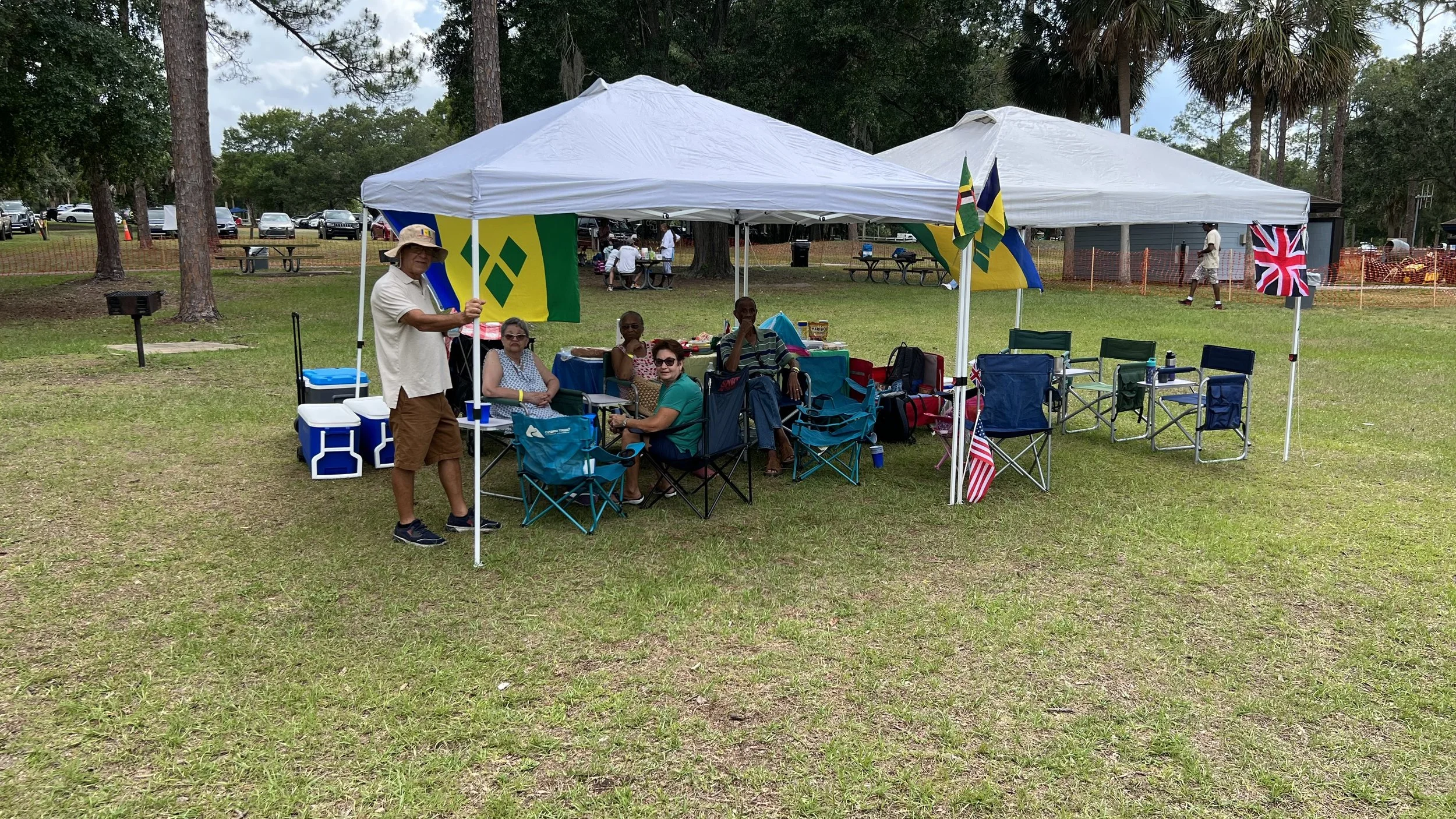 Group of people seated under a white canopy at an outdoor park, with flags from different countries hanging on the tent. Some people are sitting in camping chairs, and there are coolers nearby.