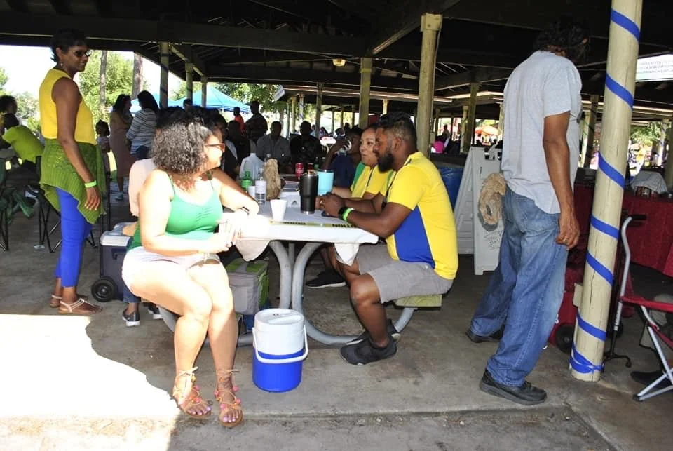 People sitting at a table under a pavilion, engaged in conversation, with others standing and walking nearby, at an outdoor social gathering or event.