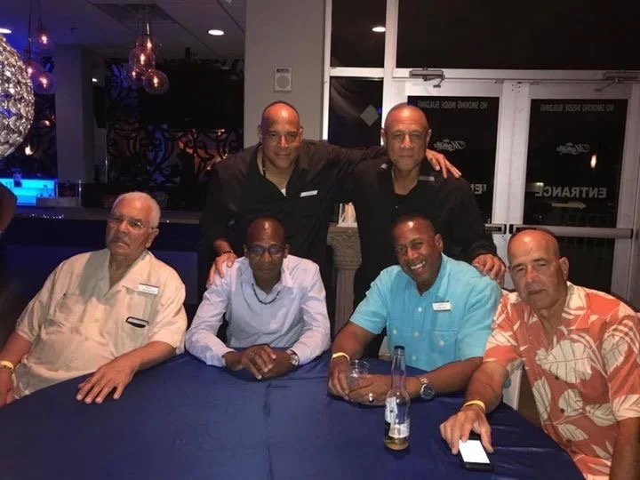 Six men sitting and standing around a table at a social gathering in a dimly lit room. Some are smiling, others with neutral expressions. One man is holding a beer bottle, and another has a smartphone on the table.