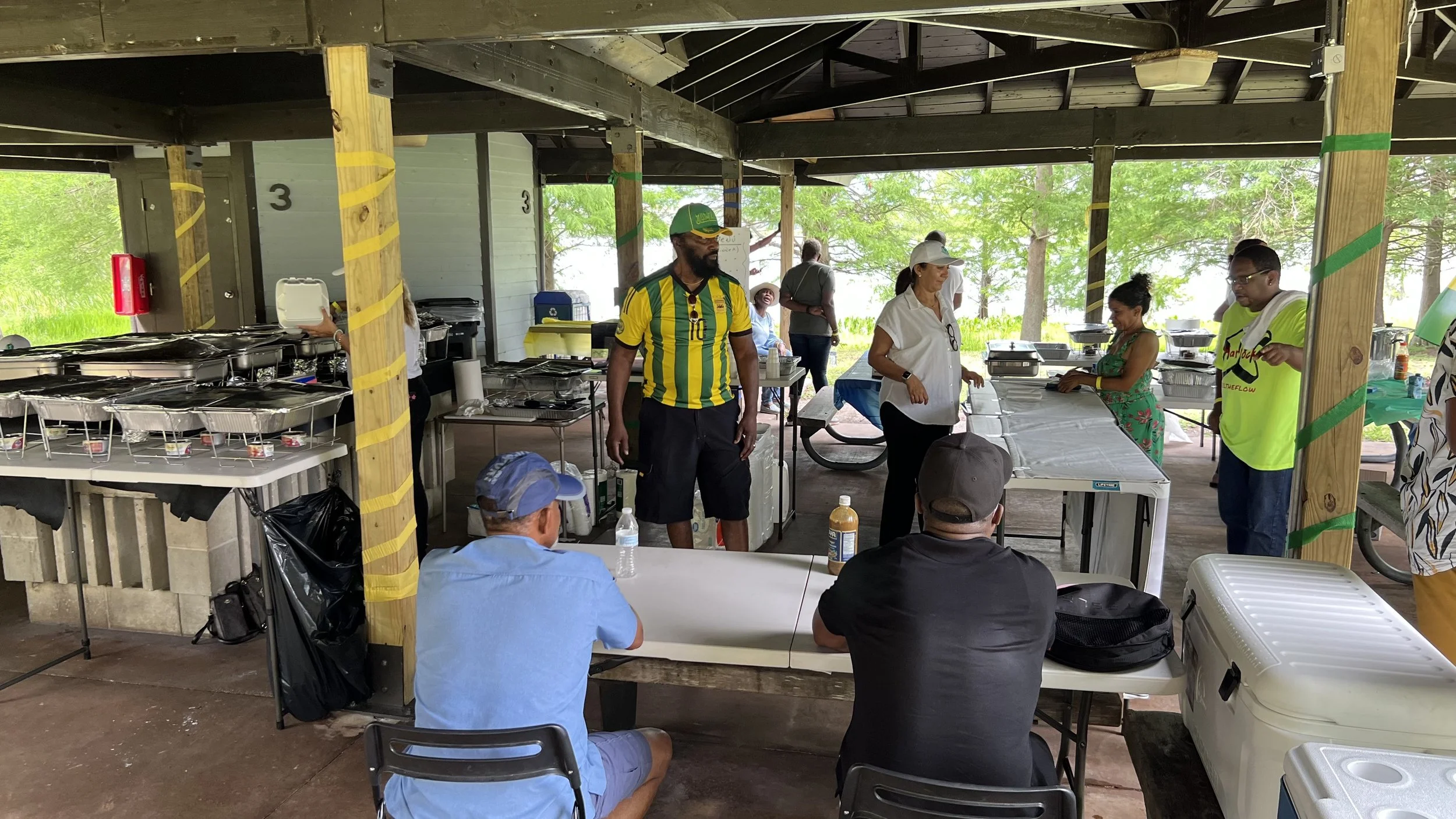 People gather under a pavilion at a park, with food stations and tables set up, some seated, others standing and serving food.