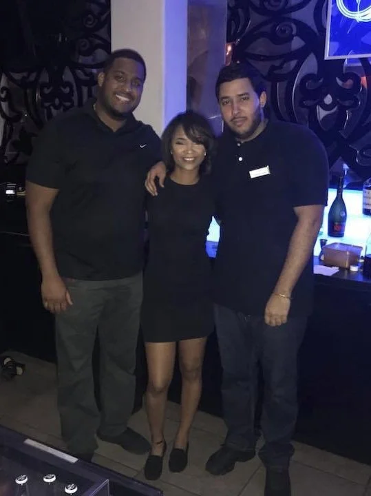 Three people standing together in a dimly lit bar or restaurant, smiling at the camera. Two men and one woman, all dressed in black, with bottles and glasses on the counter behind them.