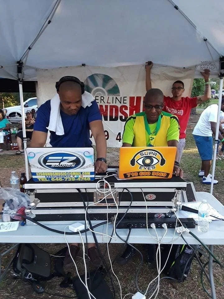 Two DJs operating turntables and equipment under a white canopy at an outdoor event, with a banner in the background.