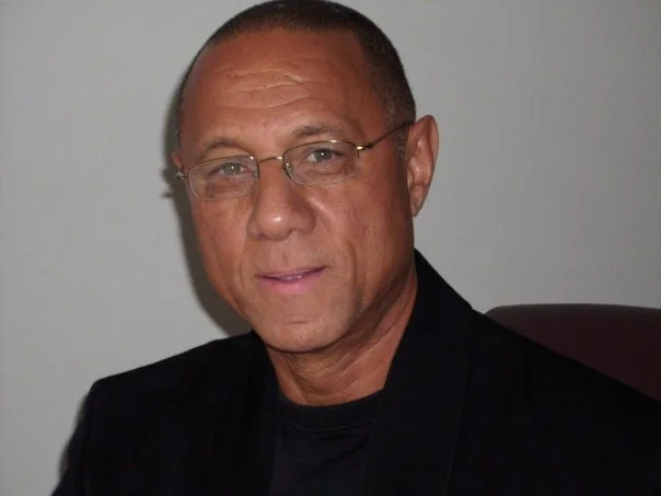 A man with glasses and short hair, wearing a dark blazer, sitting against a plain background.