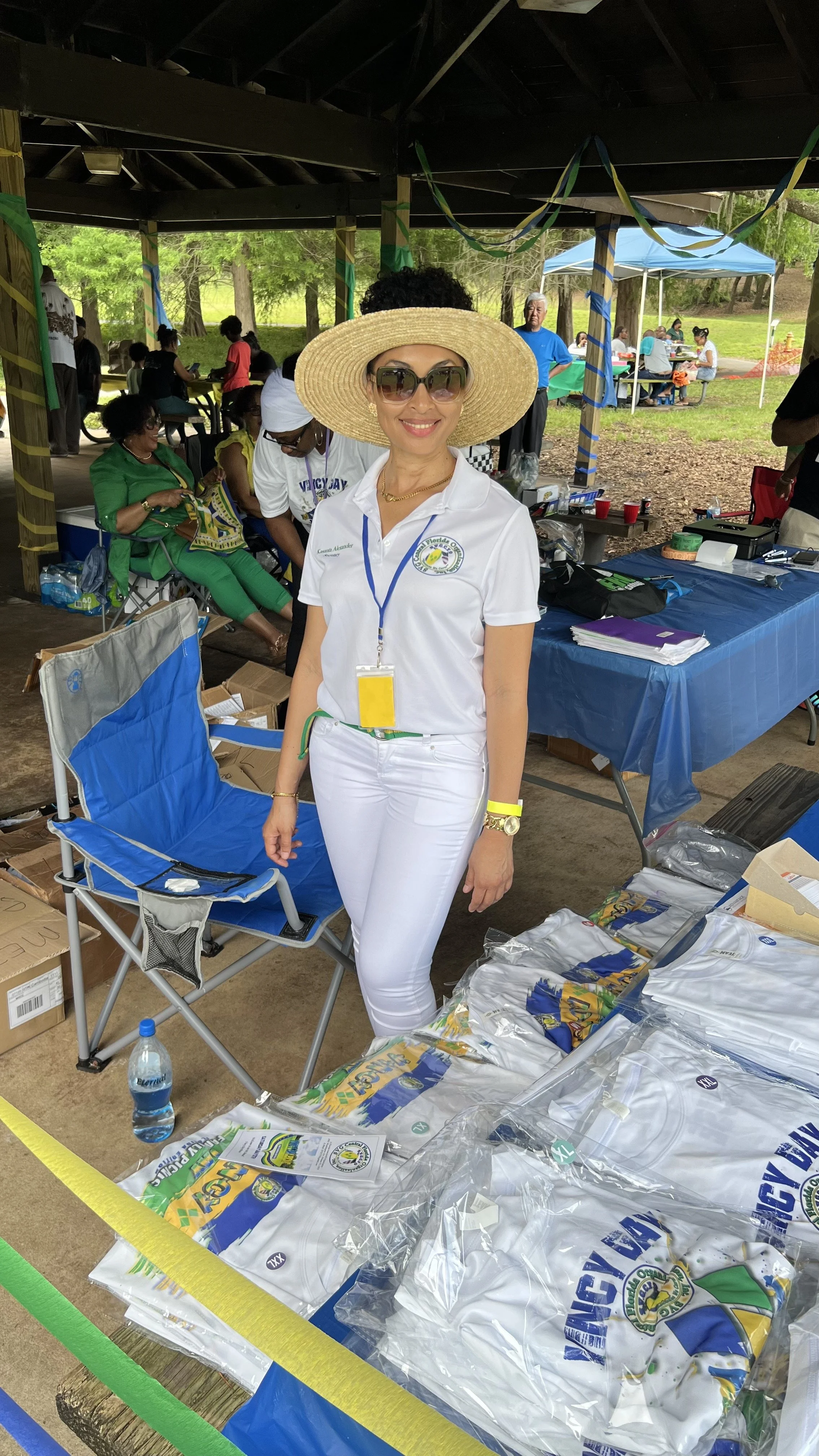 A woman wearing sunglasses, a wide-brimmed straw hat, a white polo shirt with a logo, and white pants, standing in a pavilion at an outdoor event with people sitting at tables in the background.