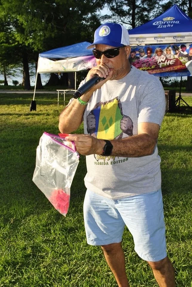 A man wearing sunglasses, a blue and white cap with a logo, a gray T-shirt, and white shorts is holding a microphone in one hand and a clear plastic bag with pink items inside in the other. He appears to be speaking or singing outdoors at a park or f