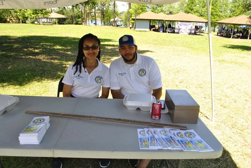 Two people sitting at a table under a canopy at an outdoor event, with brochures, a ruler, a takeout container, and a soda can on the table. Both are wearing white polo shirts with a logo, and the setting includes grassy area, trees, and other event 
