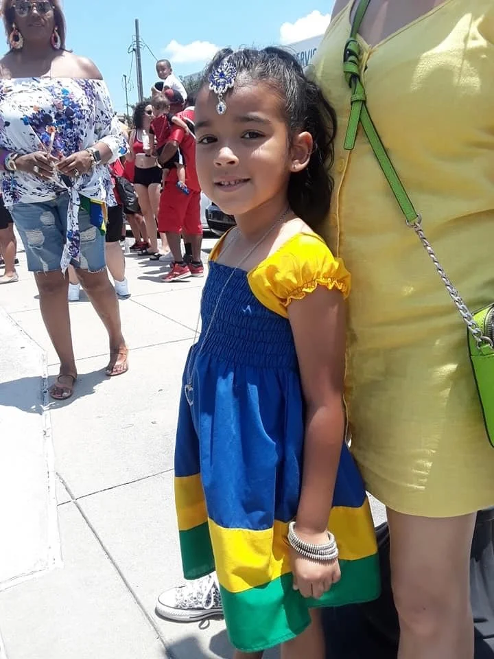 A young girl wearing a colorful dress with yellow, blue, and green sections, smiling outdoors amid a crowd, with people in casual summer clothing and a bright sky.