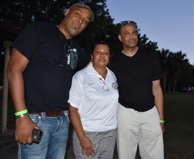 Three people standing outdoors in the evening, smiling at the camera. The man on the left is wearing a black t-shirt and holding a phone, the woman in the middle is wearing a white shirt with glasses hanging around her neck, and the man on the right 