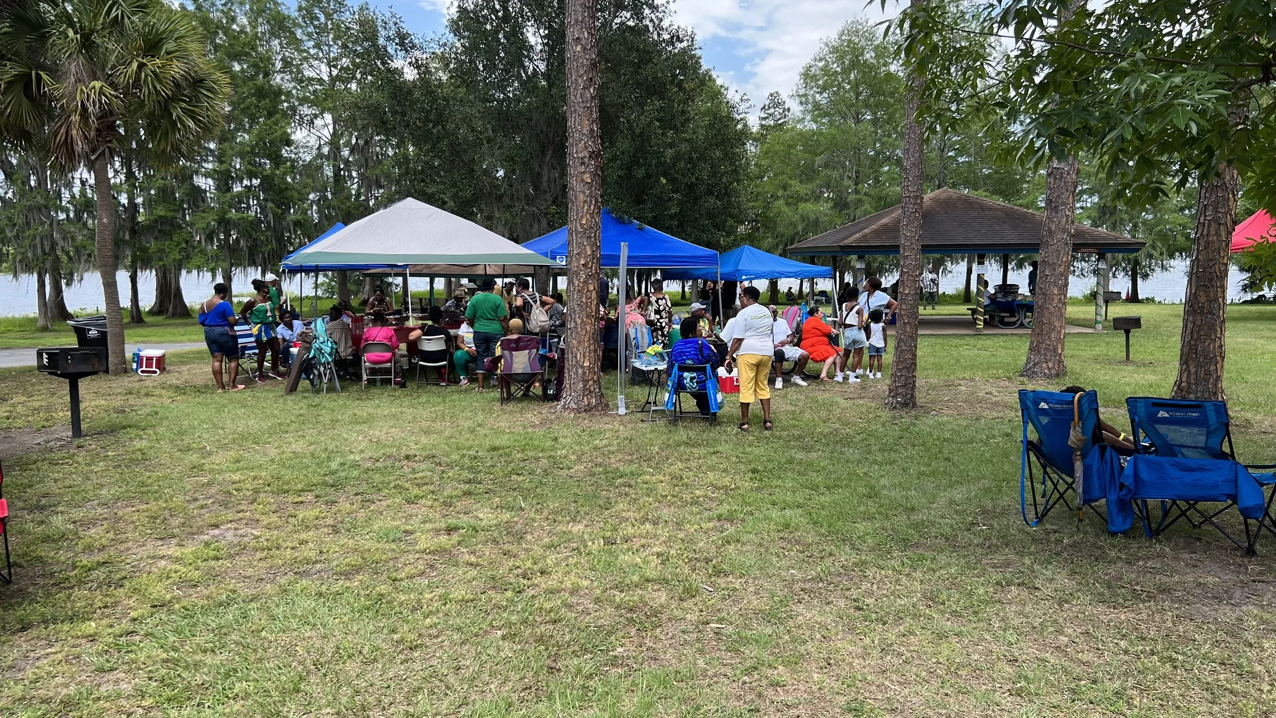 A group of people gathered at a picnic by a lakeside, with picnic tents, chairs, a pavilion, and trees surrounding the area.