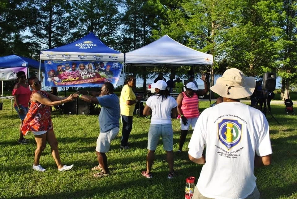 People dancing and socializing outdoors at a festival with a stage and tents in the background.