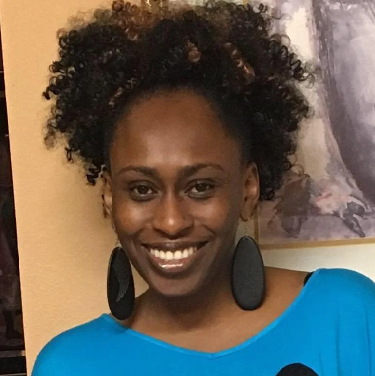 A woman with dark skin, curly hair, and a bright smile, wearing a blue top and large black oval earrings, standing indoors against a beige wall with a painting in the background.