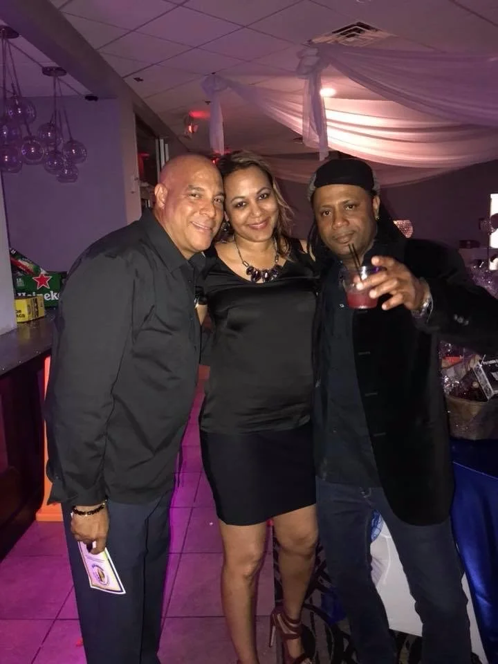Three people, two men and one woman, posing together at a party or celebration, with a bar area in the background.